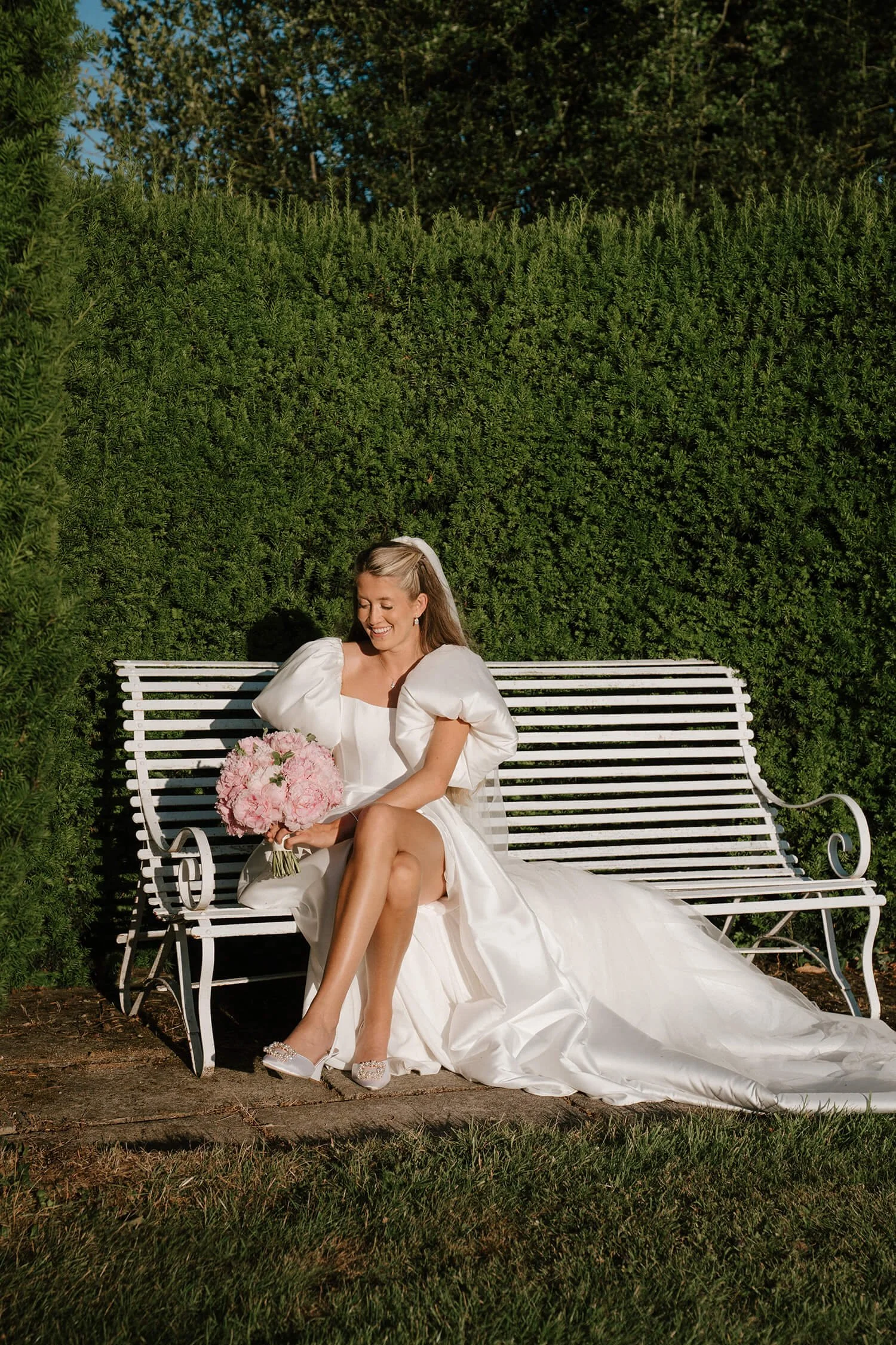 A bride in a white wedding dress sitting on a white park bench holding a bouquet of pink flowers, smiling, outdoors against a background of tall green hedges and trees.