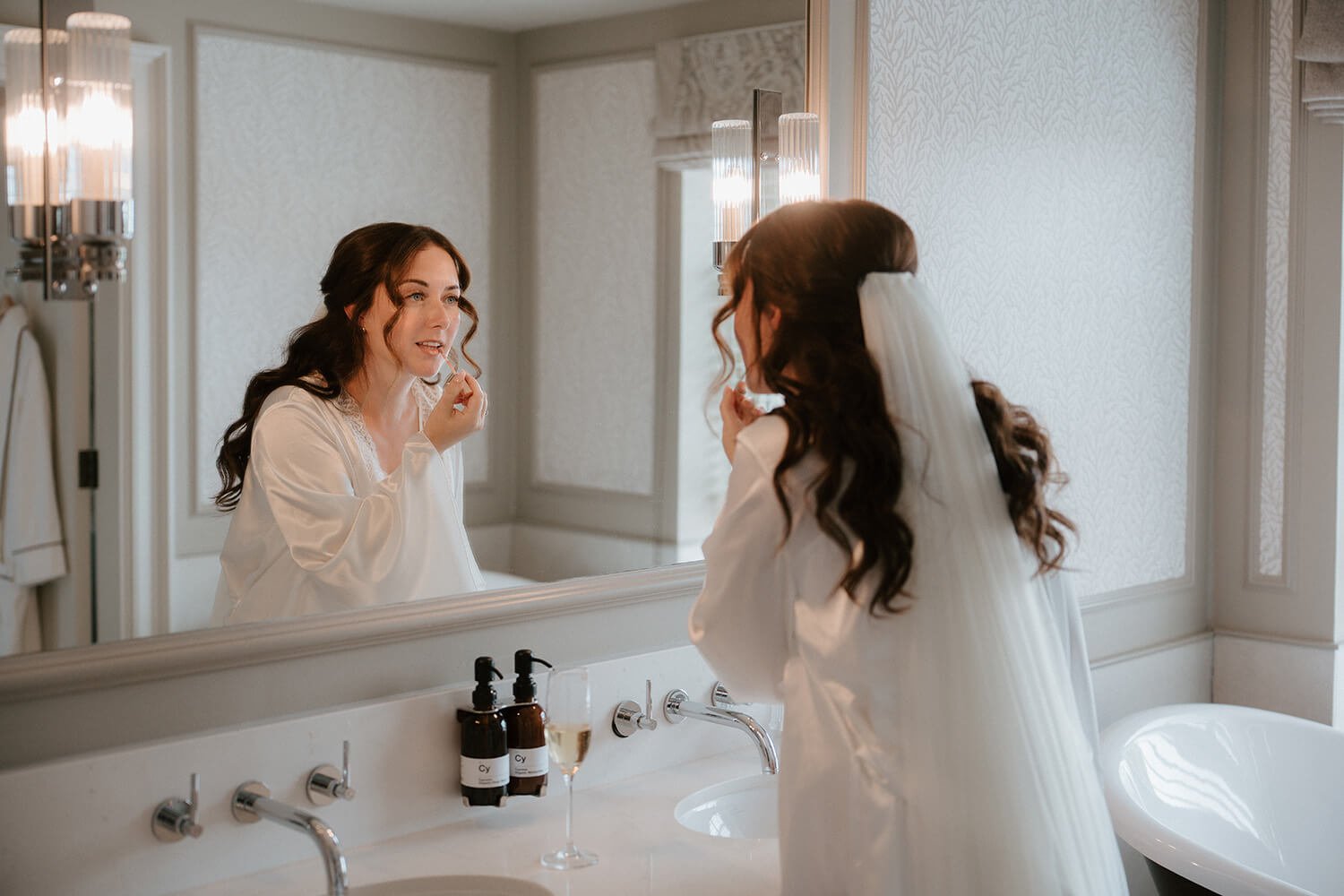 A woman, dressed as a bride in a white robe with a veil, applies makeup while looking in a bathroom mirror.