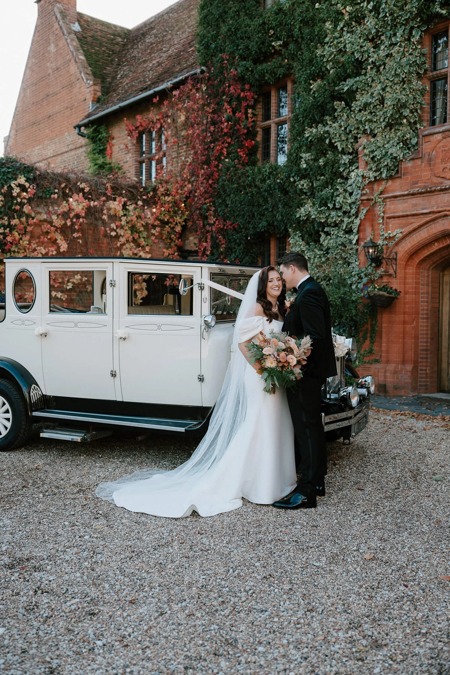 A bride and groom in wedding attire standing close and smiling in front of a vintage white car with a brick building with plants and ivy in the background.