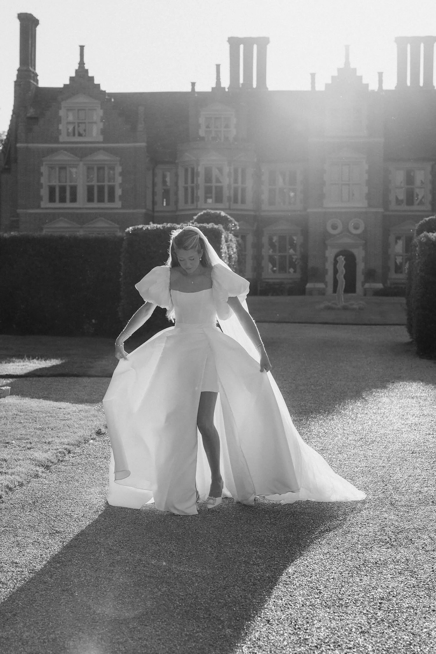 A woman in a wedding dress walking on a path in front of a large mansion, with sunlight coming from behind, casting a shadow.