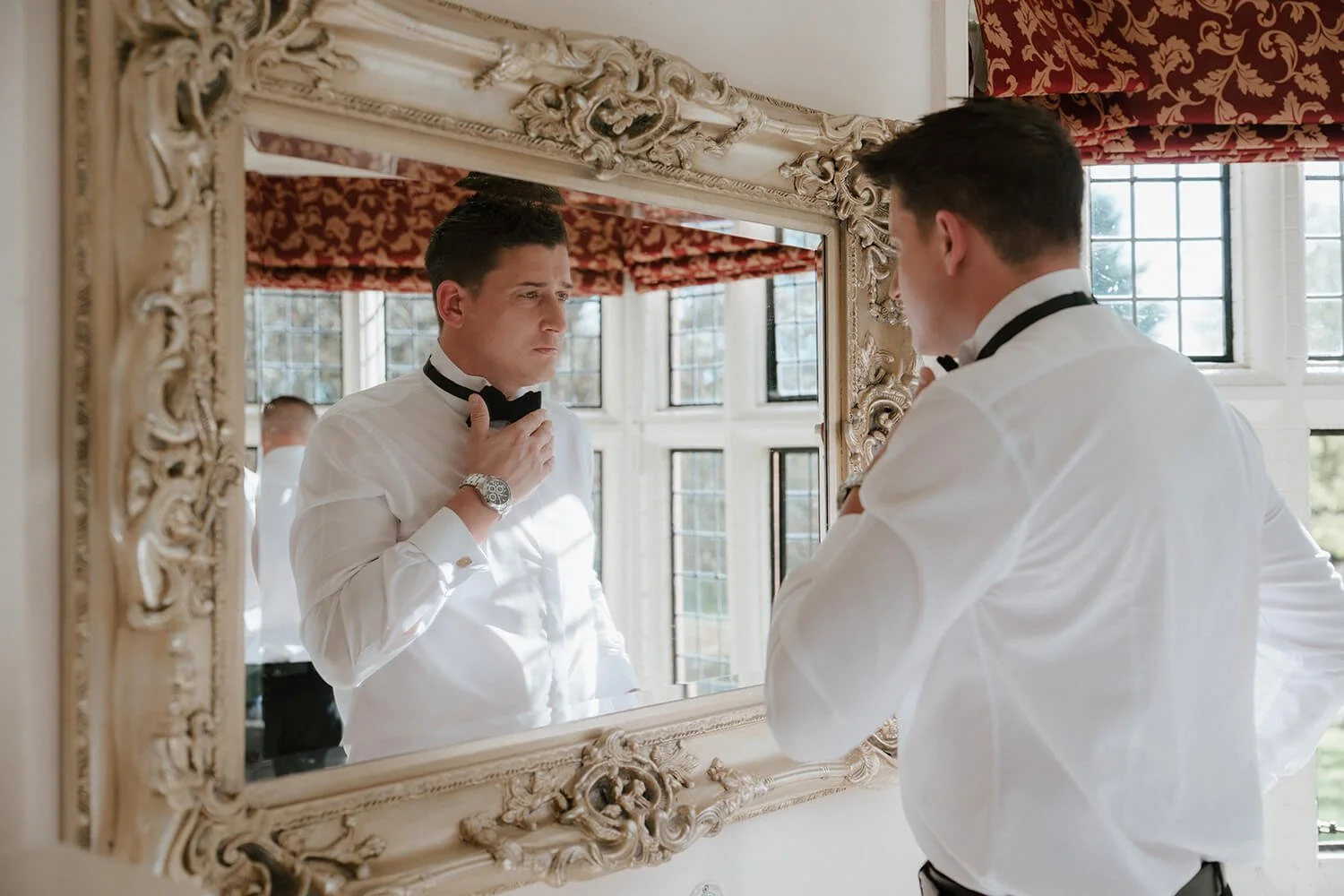 A man in a white tuxedo adjusting his black bow tie while looking at his reflection in an ornate mirror. The background includes large windows with grid panes and decorative red curtains with gold patterns.