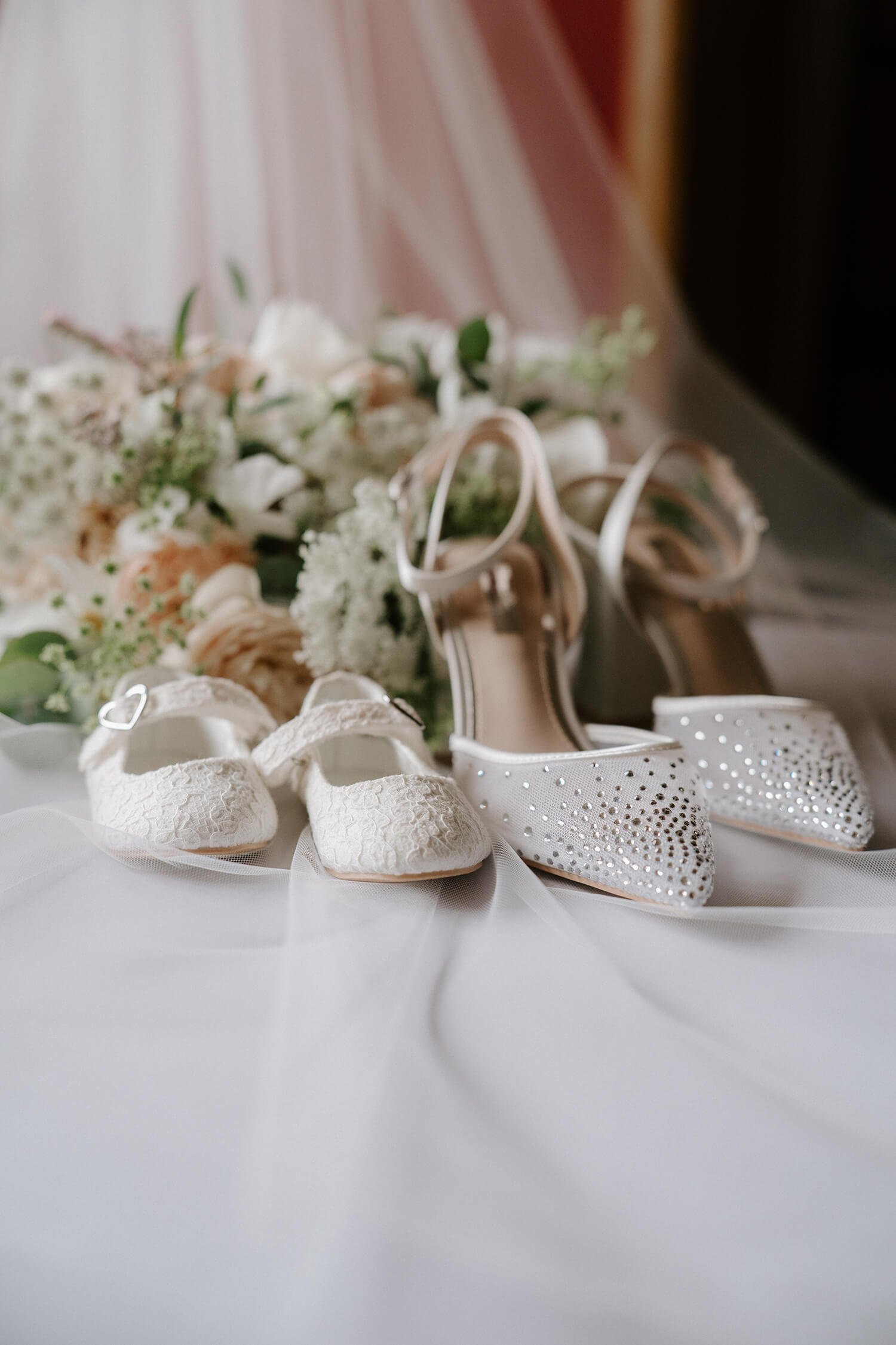 Close-up of two pairs of white wedding shoes, one with lace and the other with rhinestones, with a bouquet of flowers in the background on a white surface.
