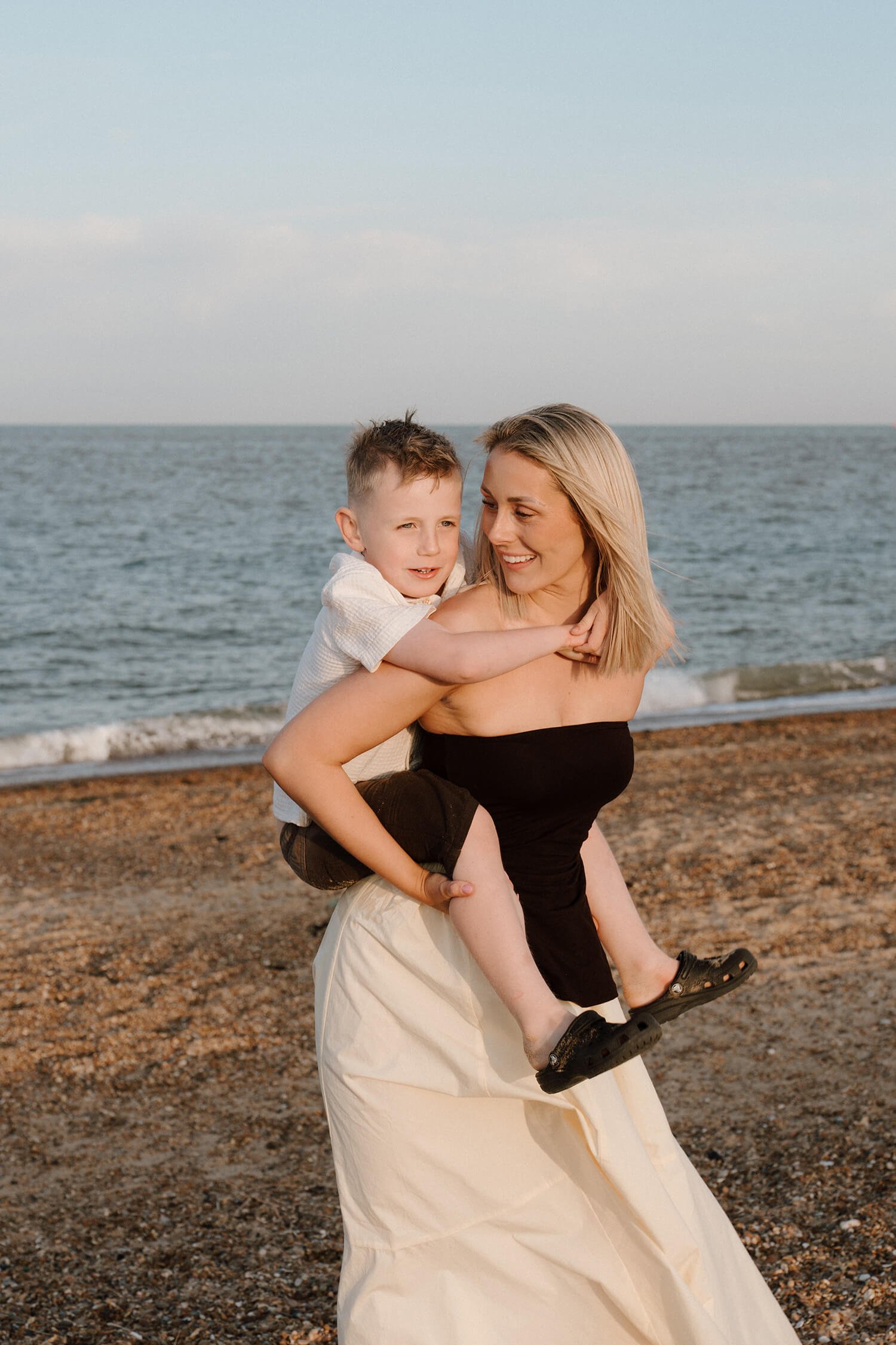 A woman carrying a young boy on her back on a beach with ocean waves in the background.
