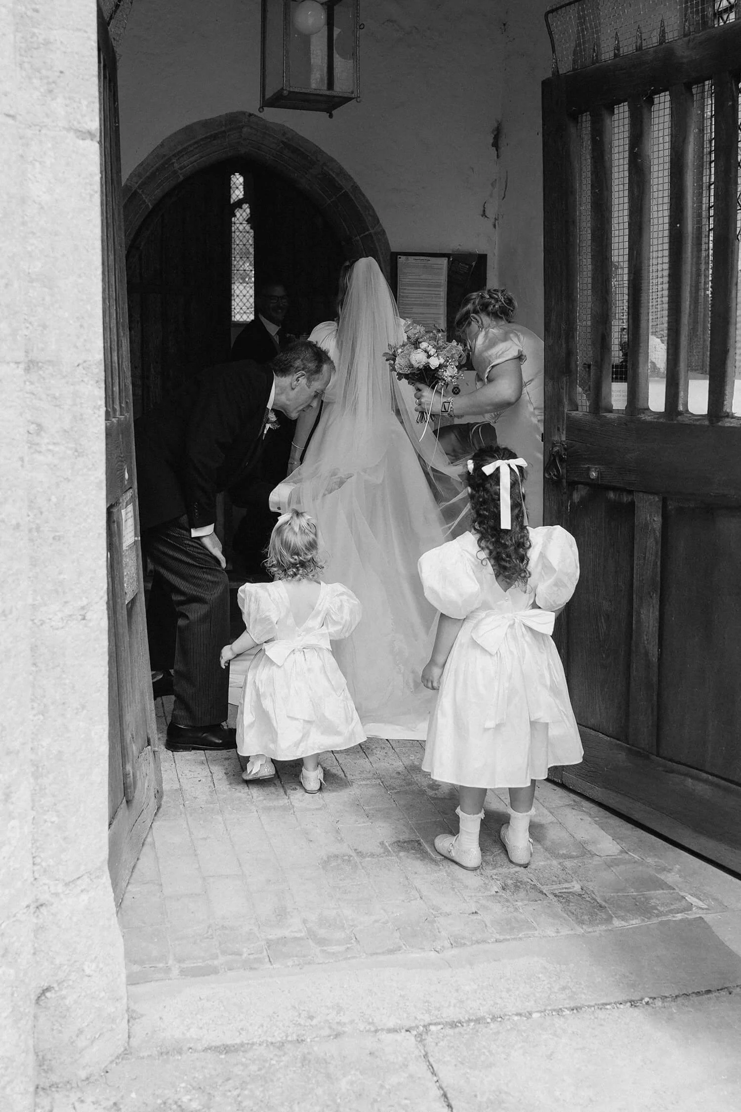 Black and white photo of a wedding scene showing a bride in a wedding dress and veil, surrounded by children in white dresses and bows, as they enter a church through an open wooden door.
