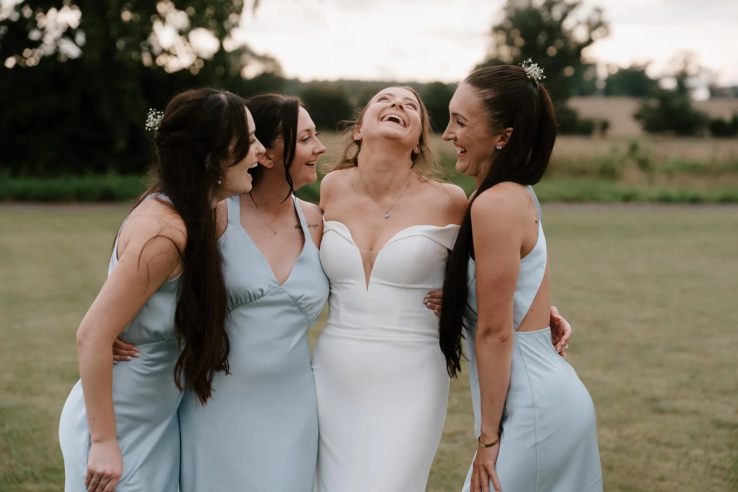 A bride in a white wedding dress laughing and smiling with her bridesmaids wearing light blue dresses outdoors during daytime.