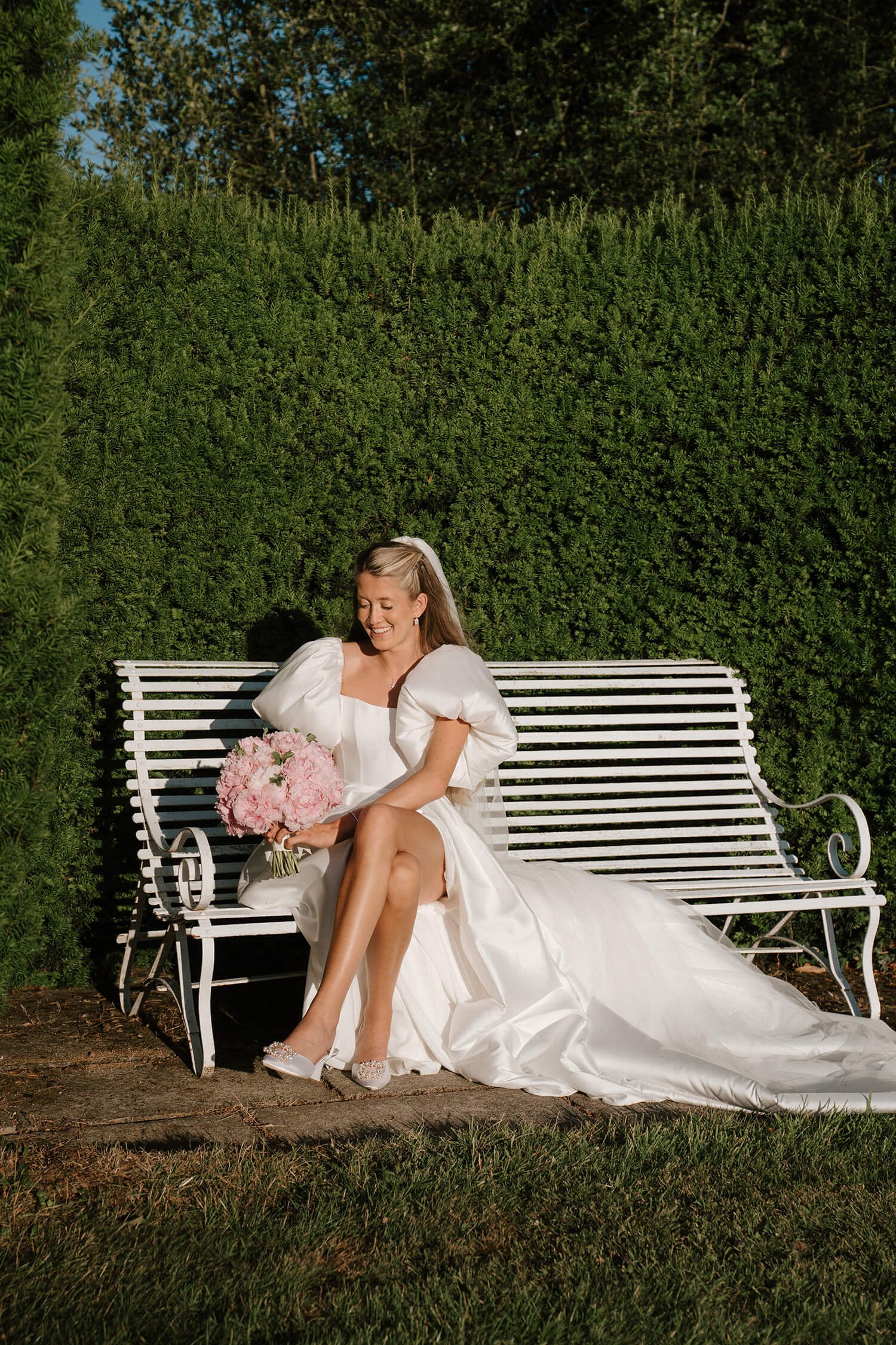 A bride sitting on a white bench in a garden, holding a bouquet of pink flowers, wearing a white wedding dress with puffy sleeves, smiling happily.