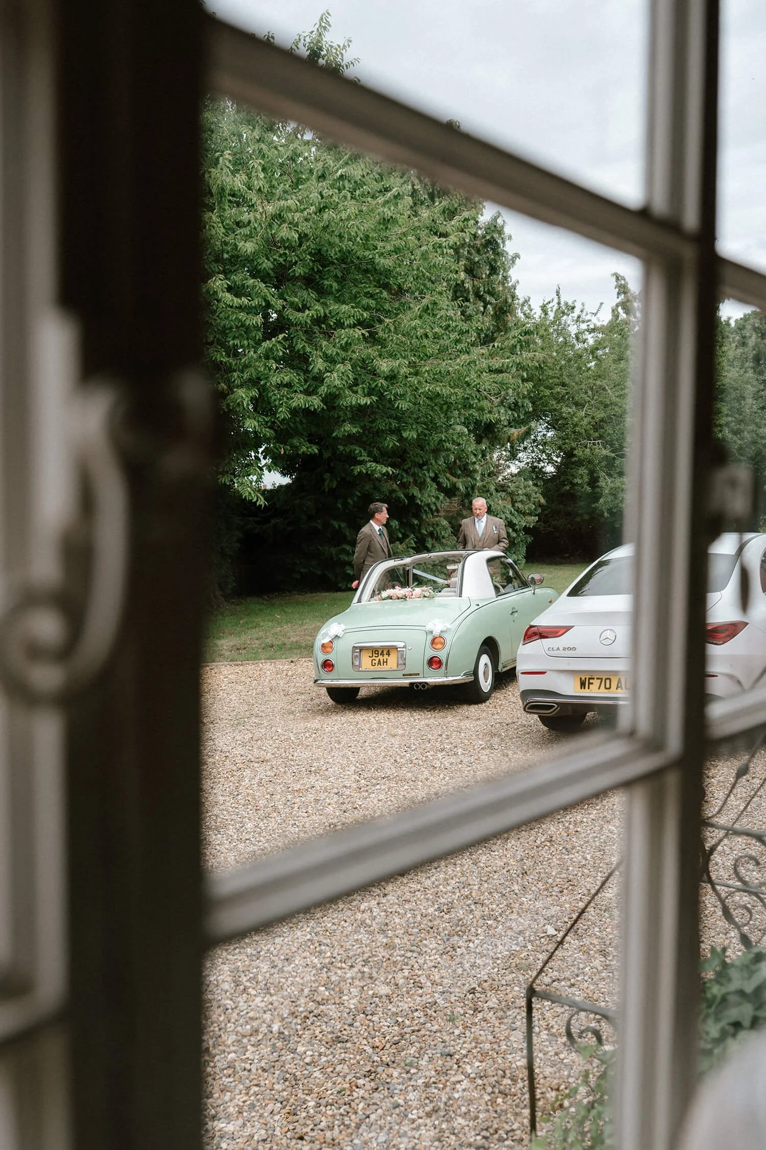 Two men in suits standing outside near a vintage green car with a flower arrangement on the trunk, viewed through a window.