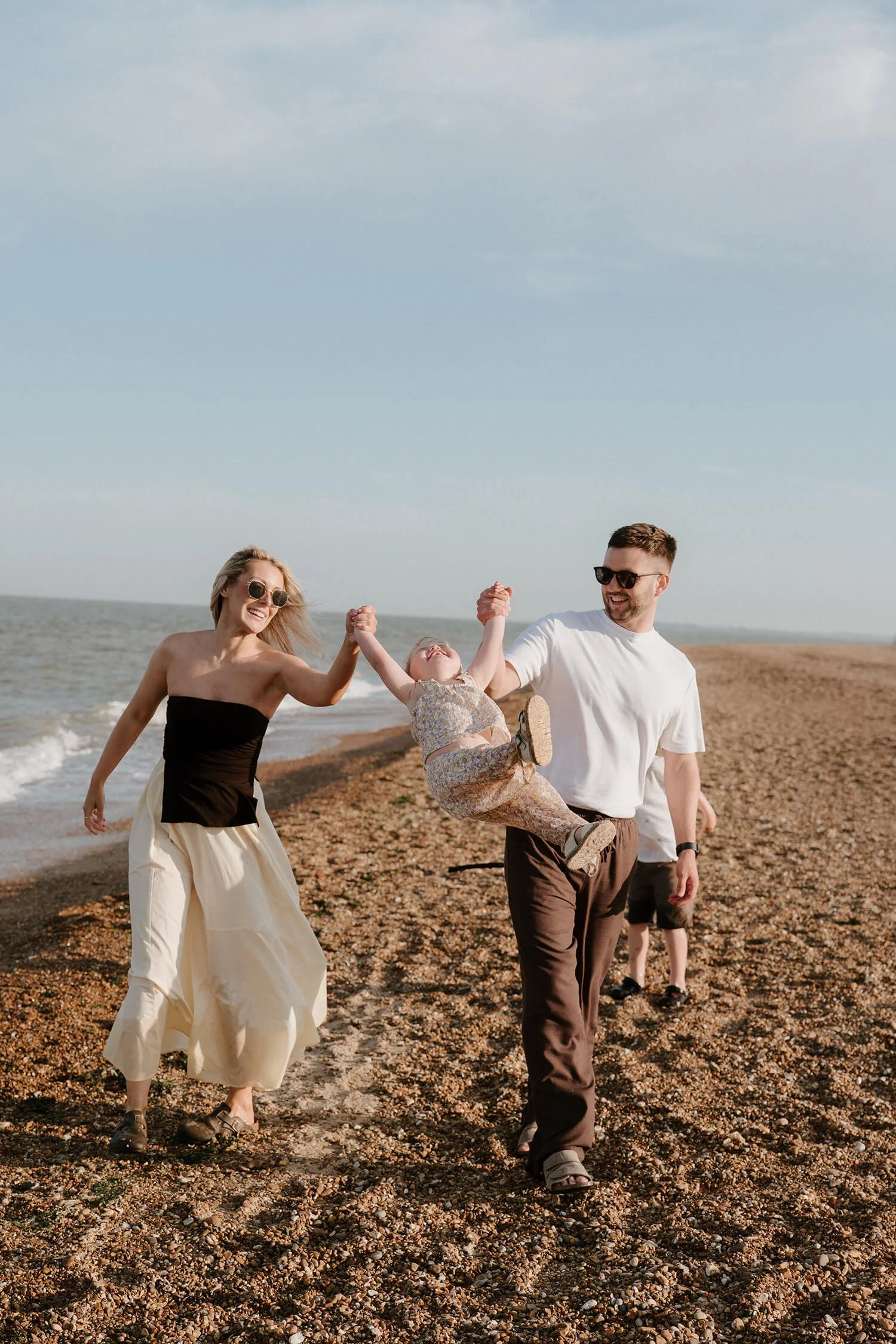 A family of three playing on a pebble beach, with the ocean in the background, during daytime. The father is holding the daughter up in the air, and the mother is holding the daughter's other hand. The family is smiling and wearing casual summer clot