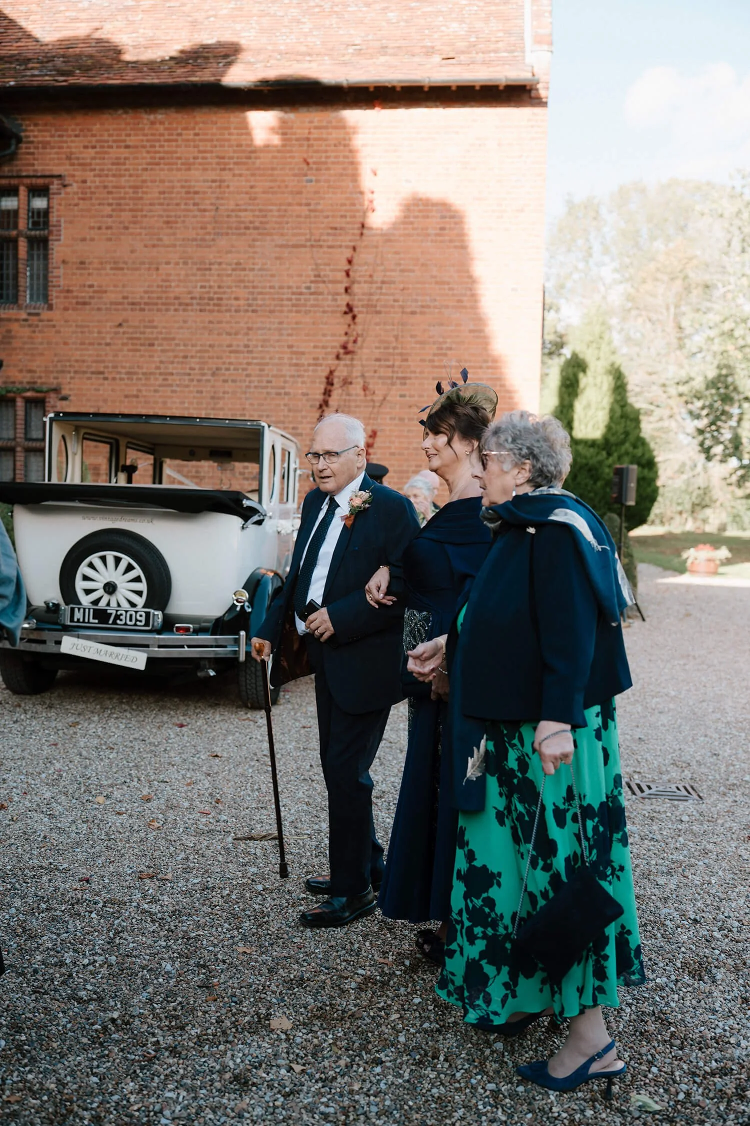 Group of elderly and middle-aged people standing outside near a vintage car with a 'Just Married' sign, dressed for a wedding celebration, with a brick building and greenery in the background.