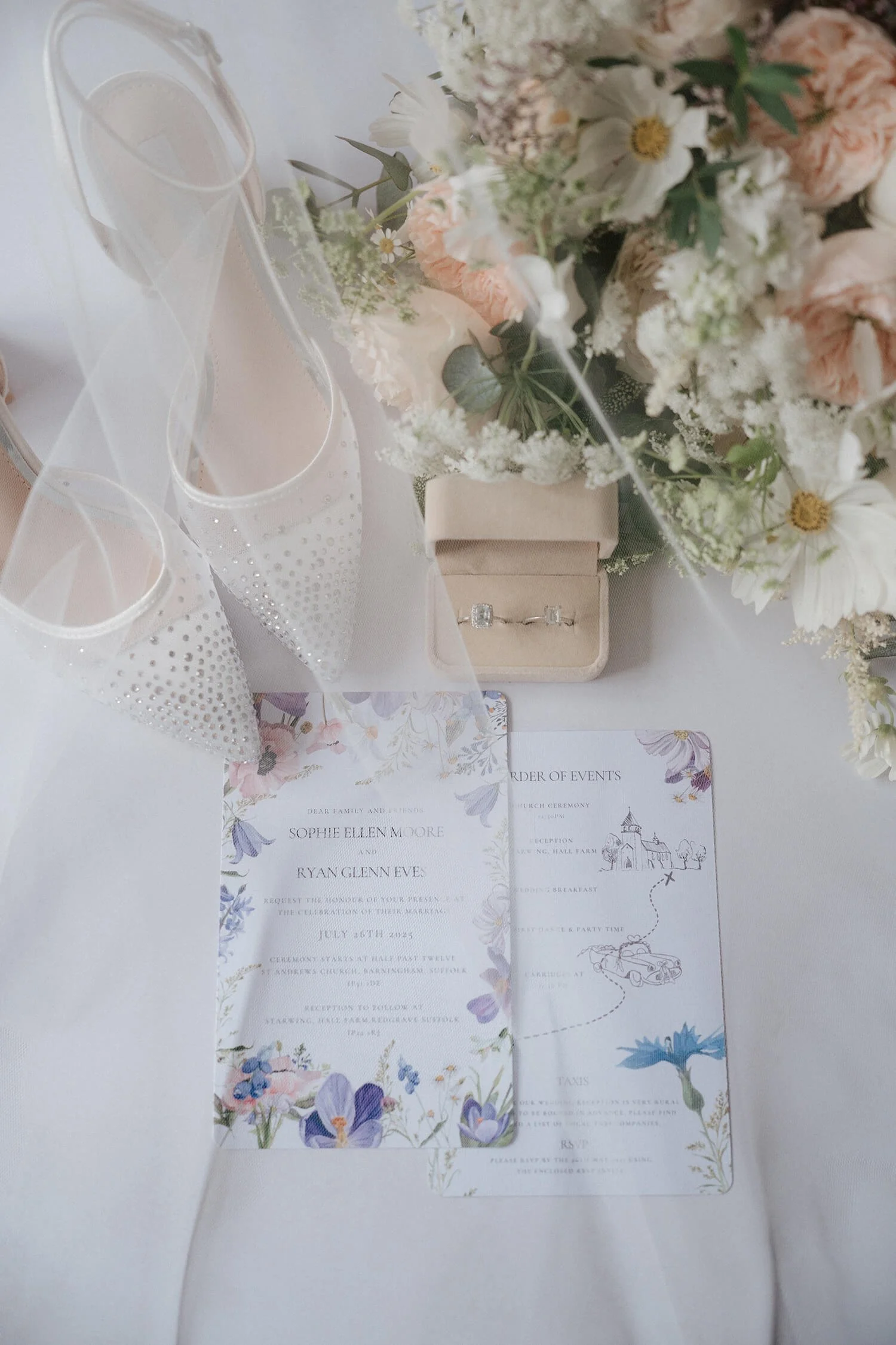 Wedding invitation with floral design, a jewelry box with rings, a bouquet of flowers, two champagne glasses with pearl accents, all on a white table.