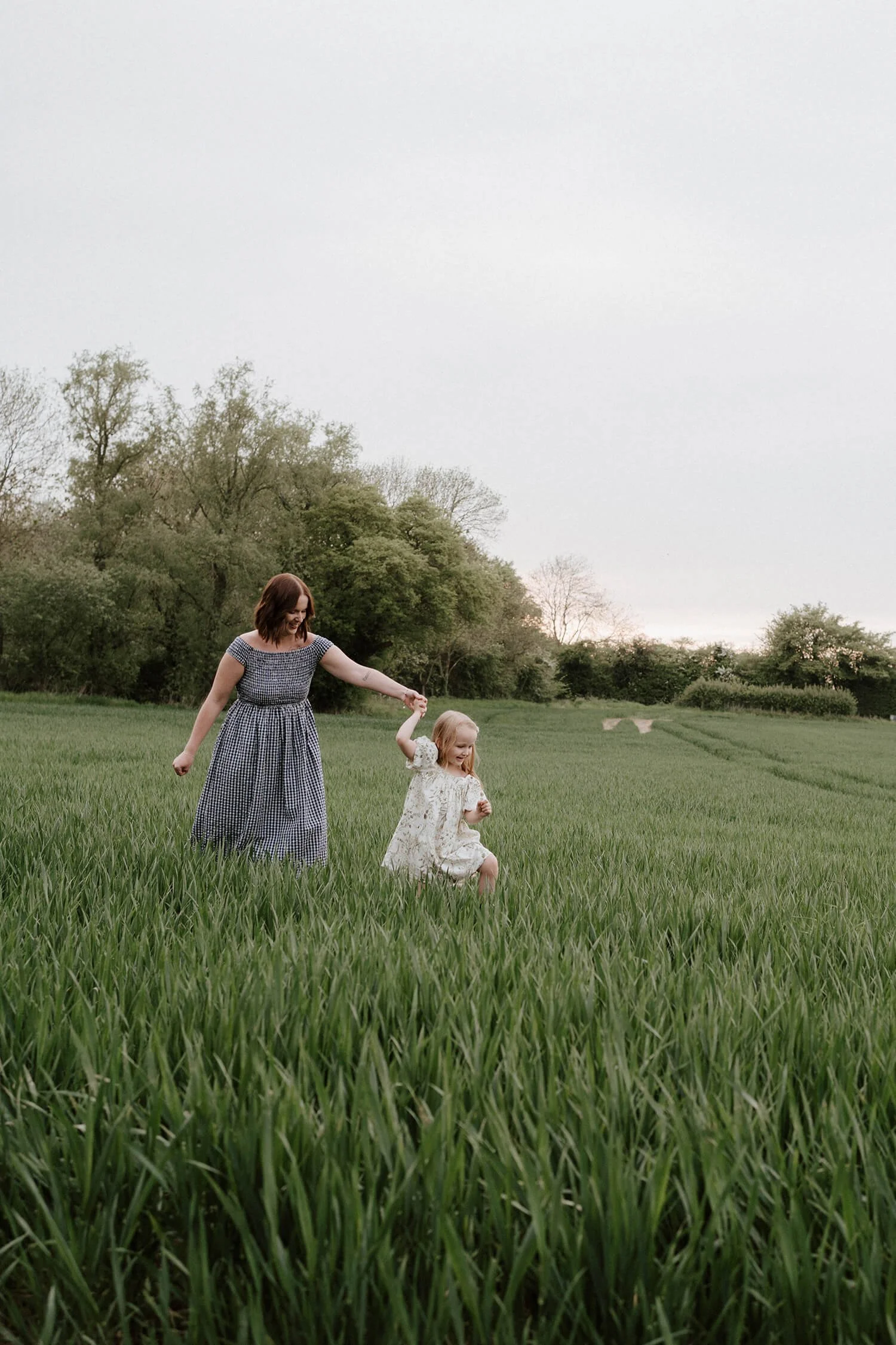 A woman and young girl playing and holding hands in a green grassy field with trees in the background, during what appears to be late afternoon or early evening.