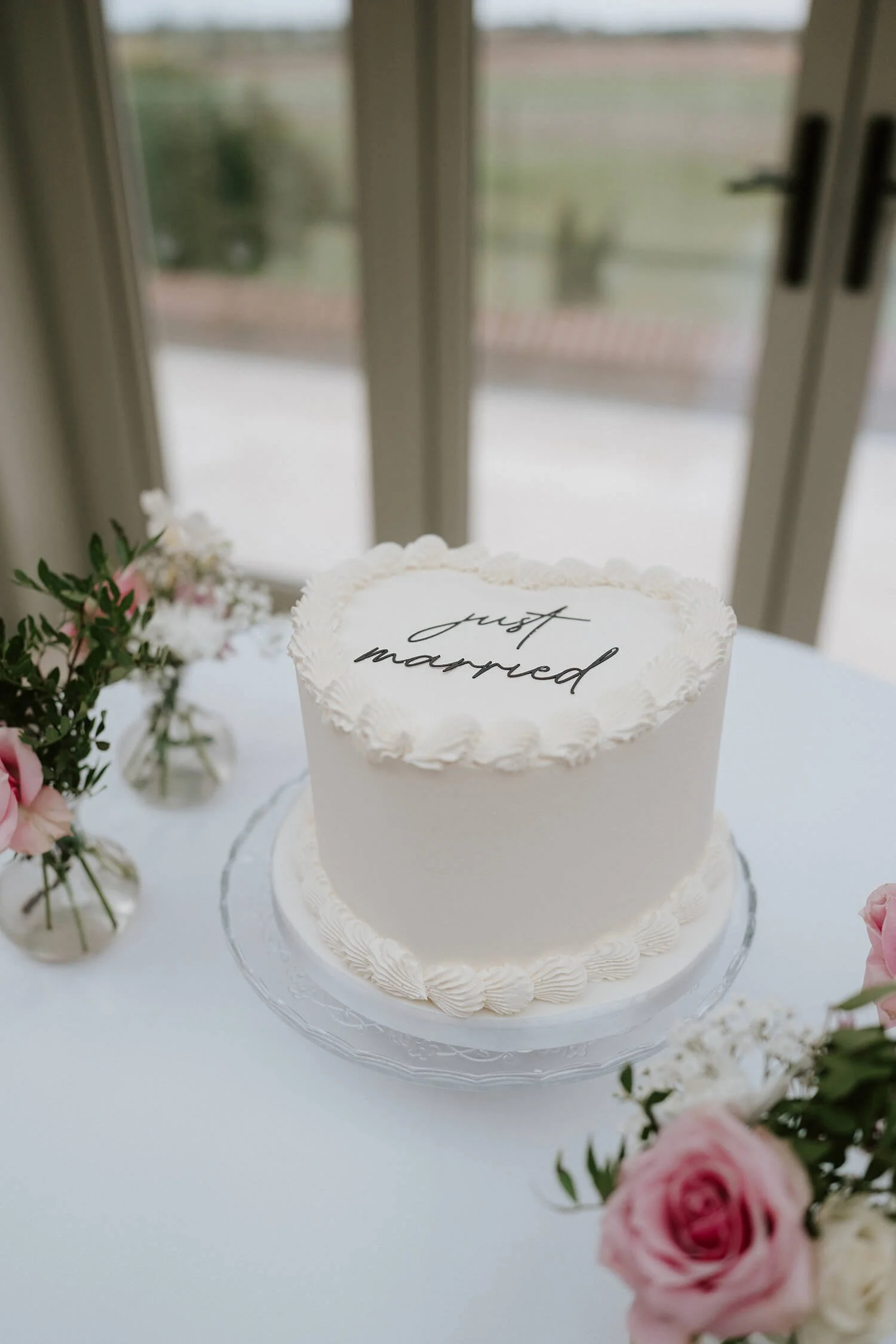 A white wedding cake with the words 'just married' written on top, surrounded by pink and white flowers, placed on a table near a glass door.