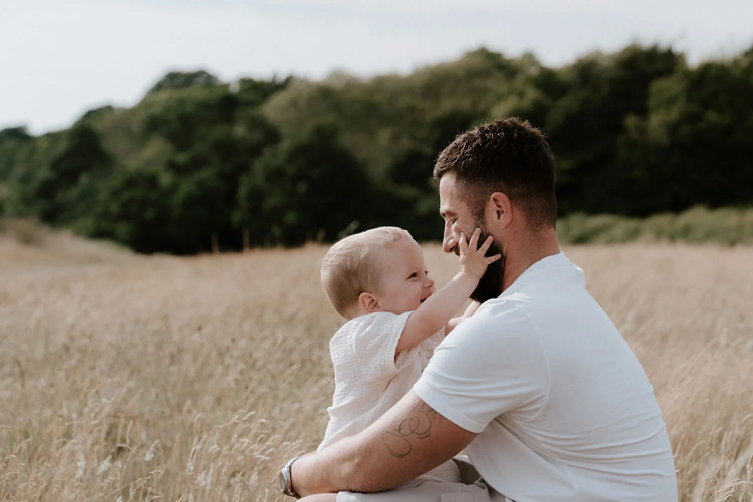 A dad and his young child smiling and touching faces in a field of tall grass with green trees in the background.