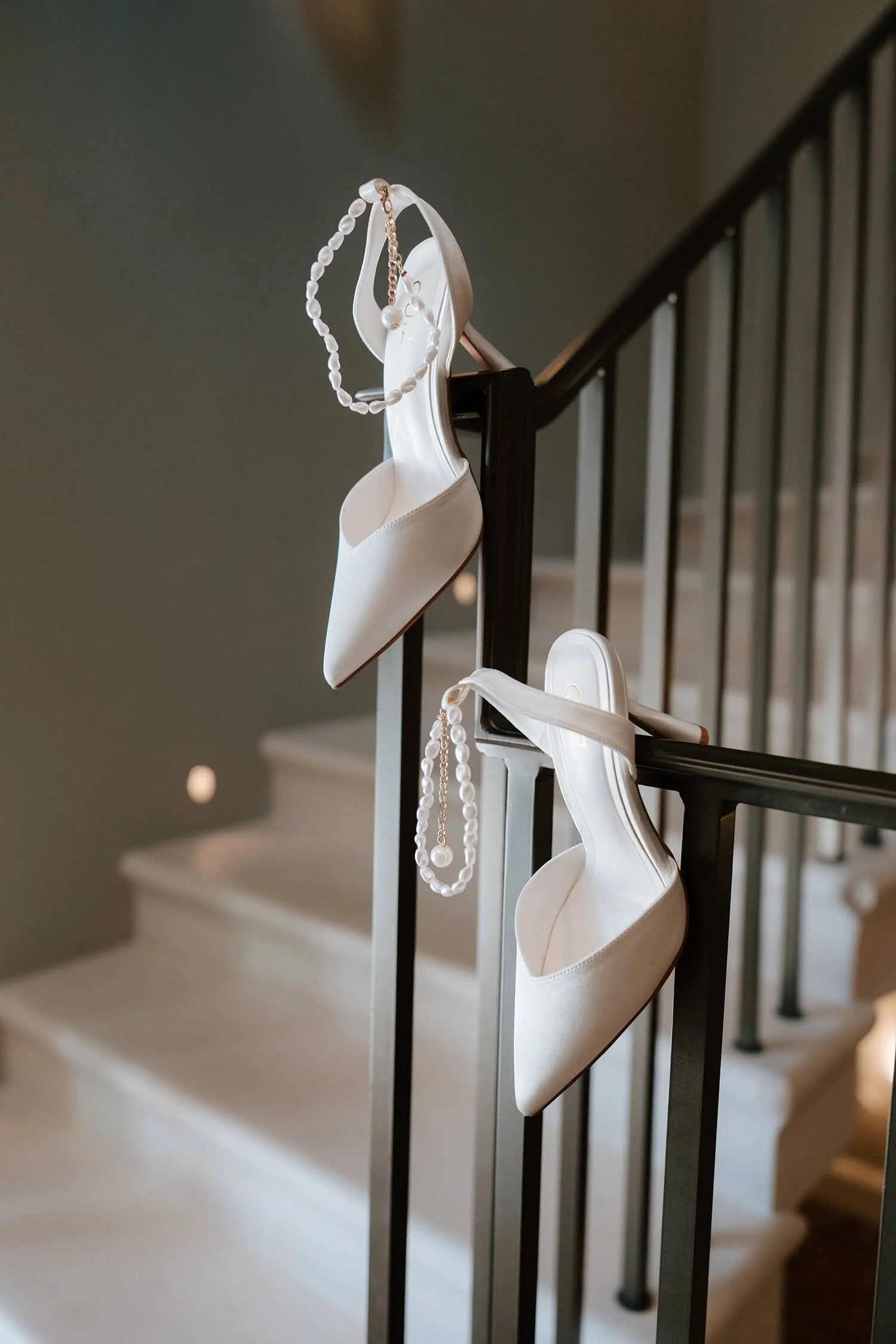 A pair of white satin high-heeled shoes hang on the black metal railing of a staircase. Two pearl necklaces with gold chains are also draped over the shoes.
