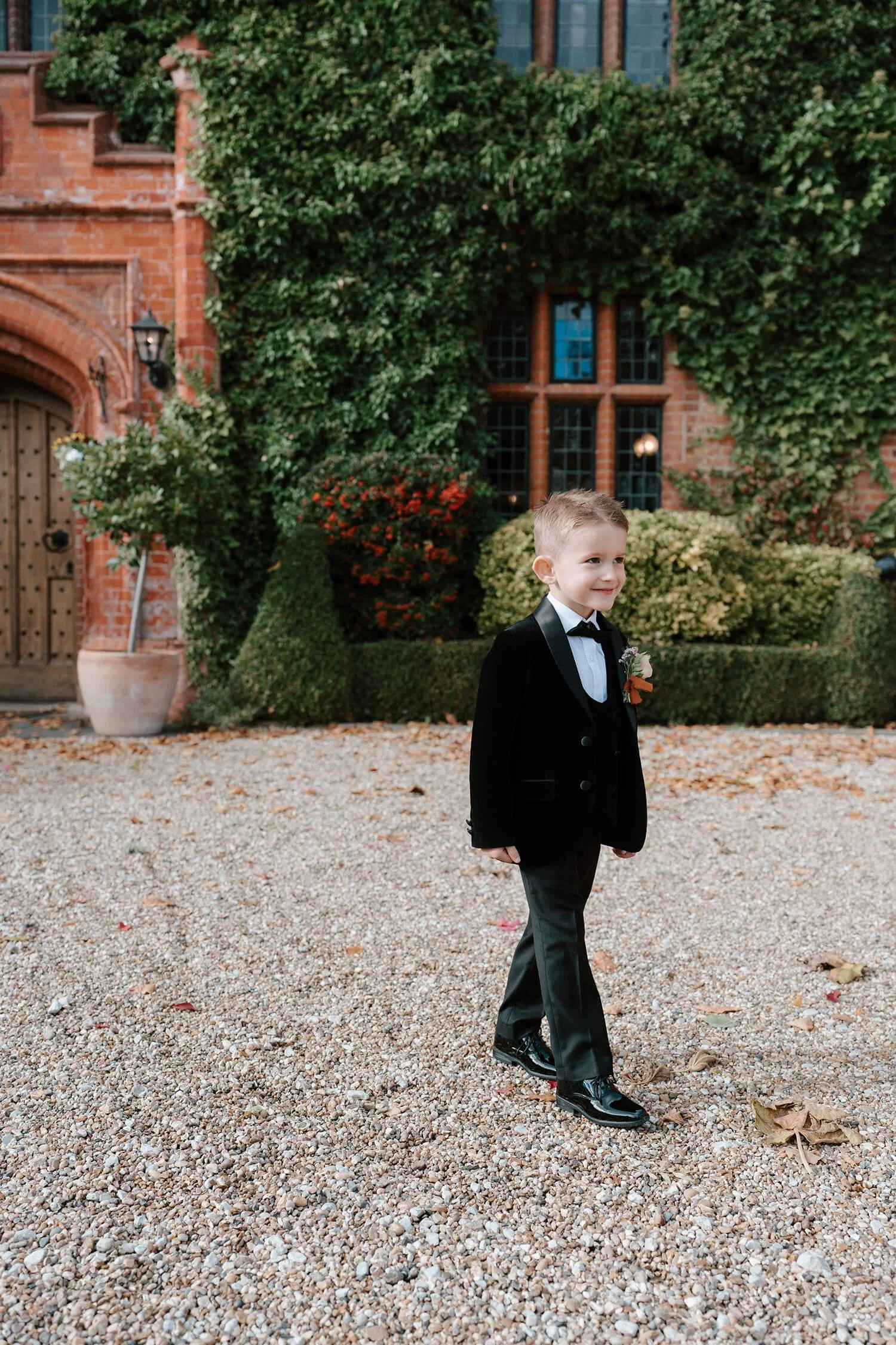 A young boy in a black tuxedo with a bow tie and boutonniere, smiling and walking outdoors on a gravel path in front of a building covered with ivy and surrounded by plants and fallen autumn leaves.
