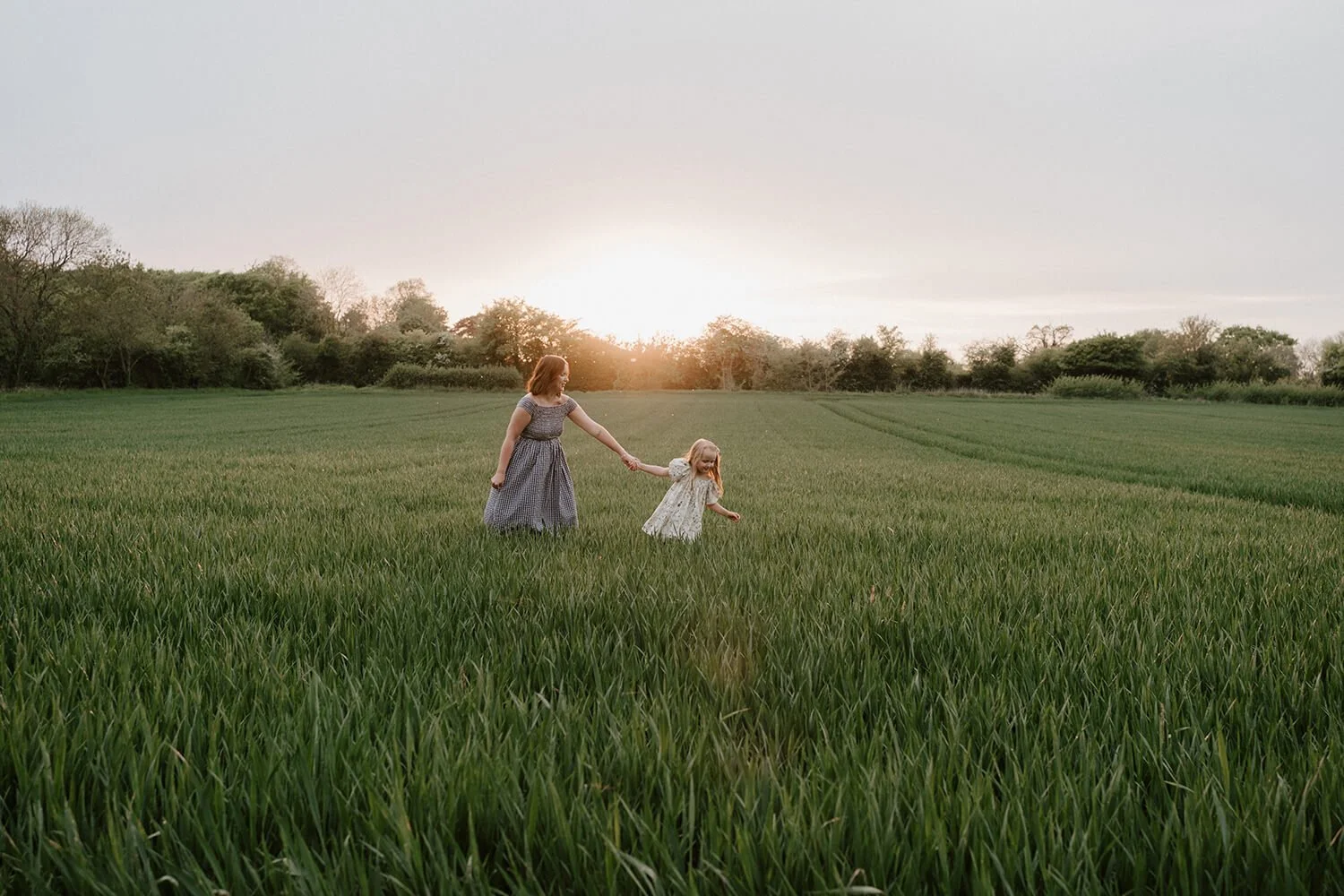 A woman and a young girl holding hands and walking through a green field at sunset, with trees in the background.