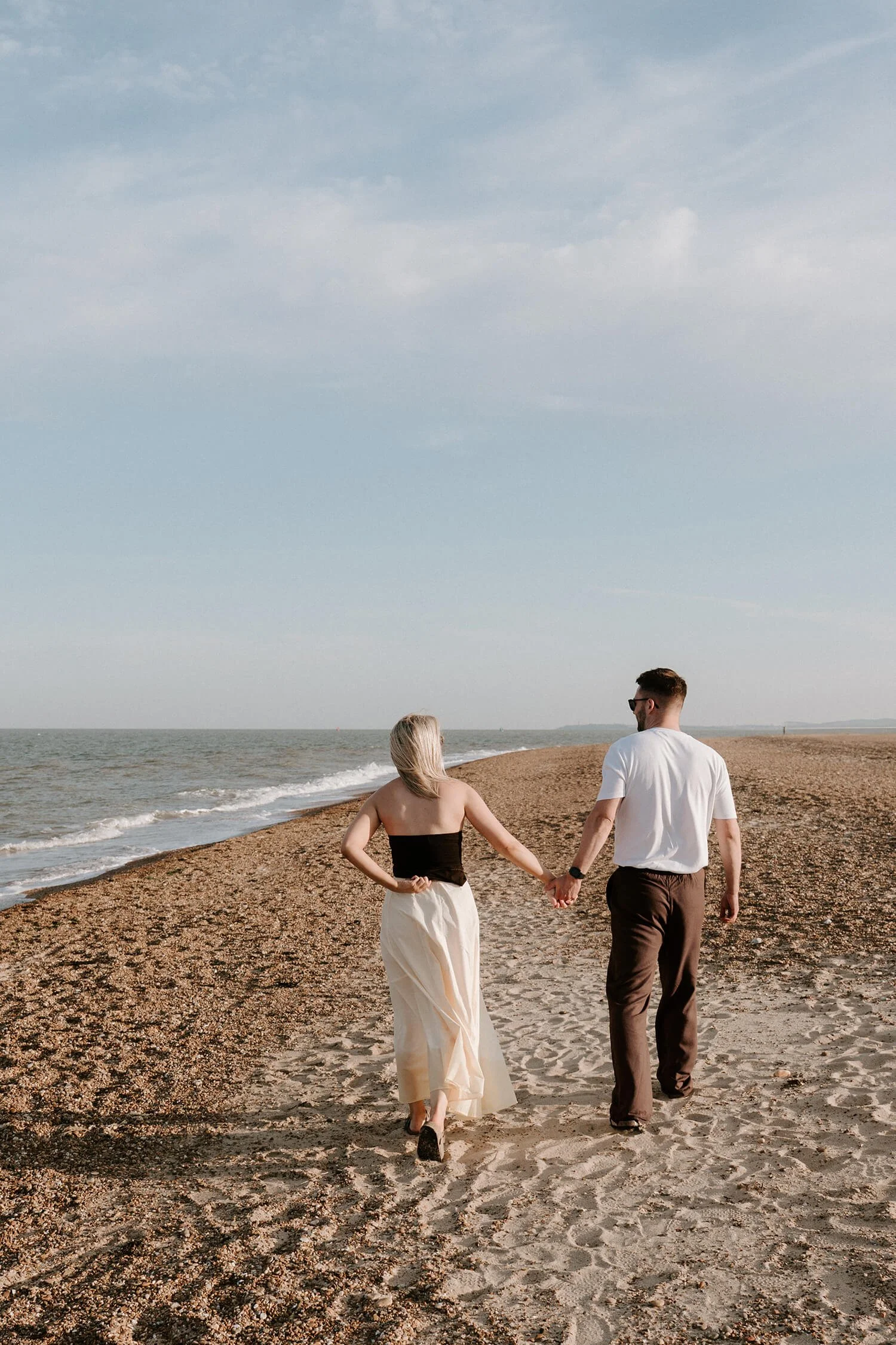 A couple holding hands and walking along the beach with the ocean on their left, under a partly cloudy sky.