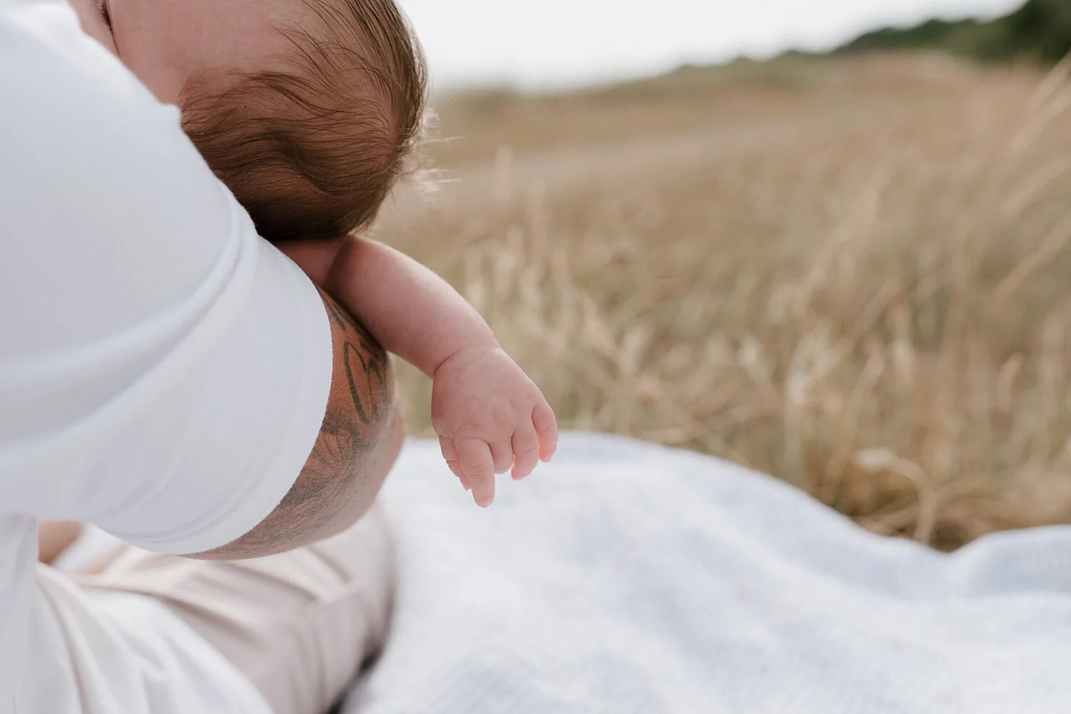 A person with a tattoo on their arm holding a baby with brown hair, outdoors in a natural setting with tall grass in the background.