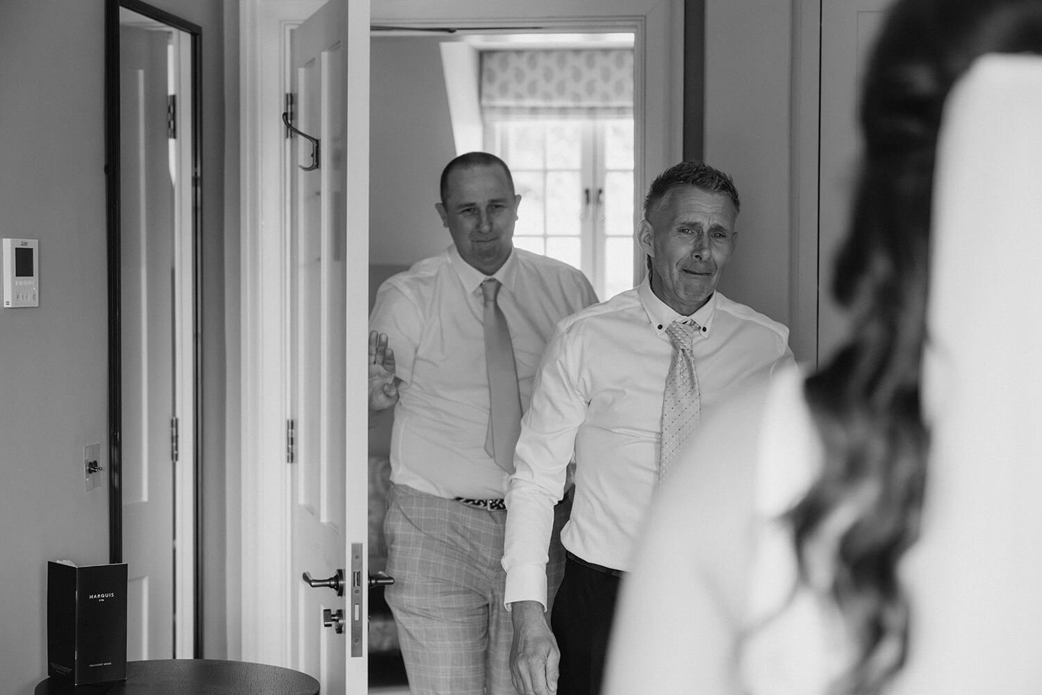 Three men in dress shirts and ties, one with a patterned tie, are standing in a doorway, appearing emotional and crying, with a woman partially visible in the foreground, likely during a wedding or emotional moment.