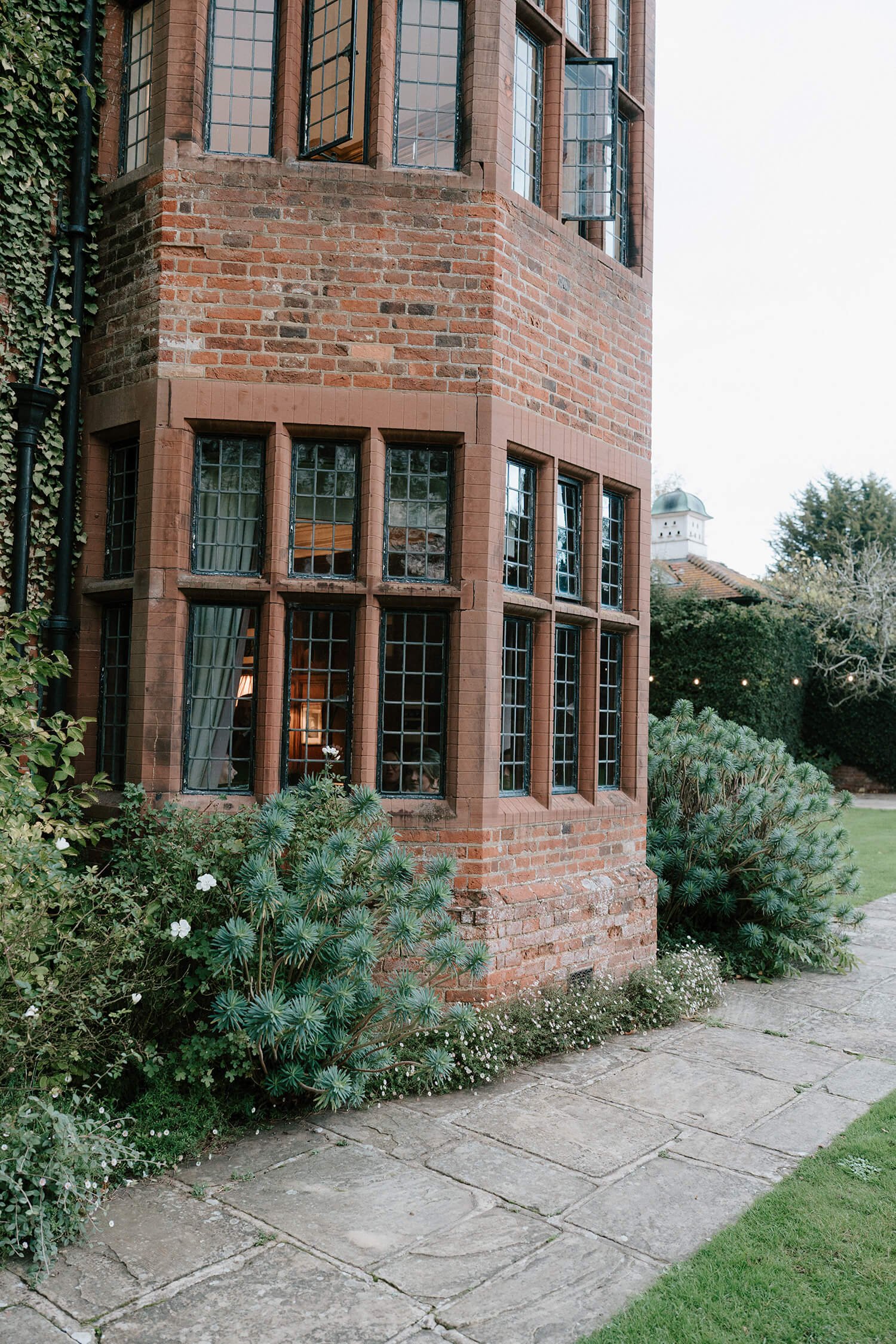 Exterior of a brick building with tall, multi-pane windows, surrounded by greenery and a stone pathway.