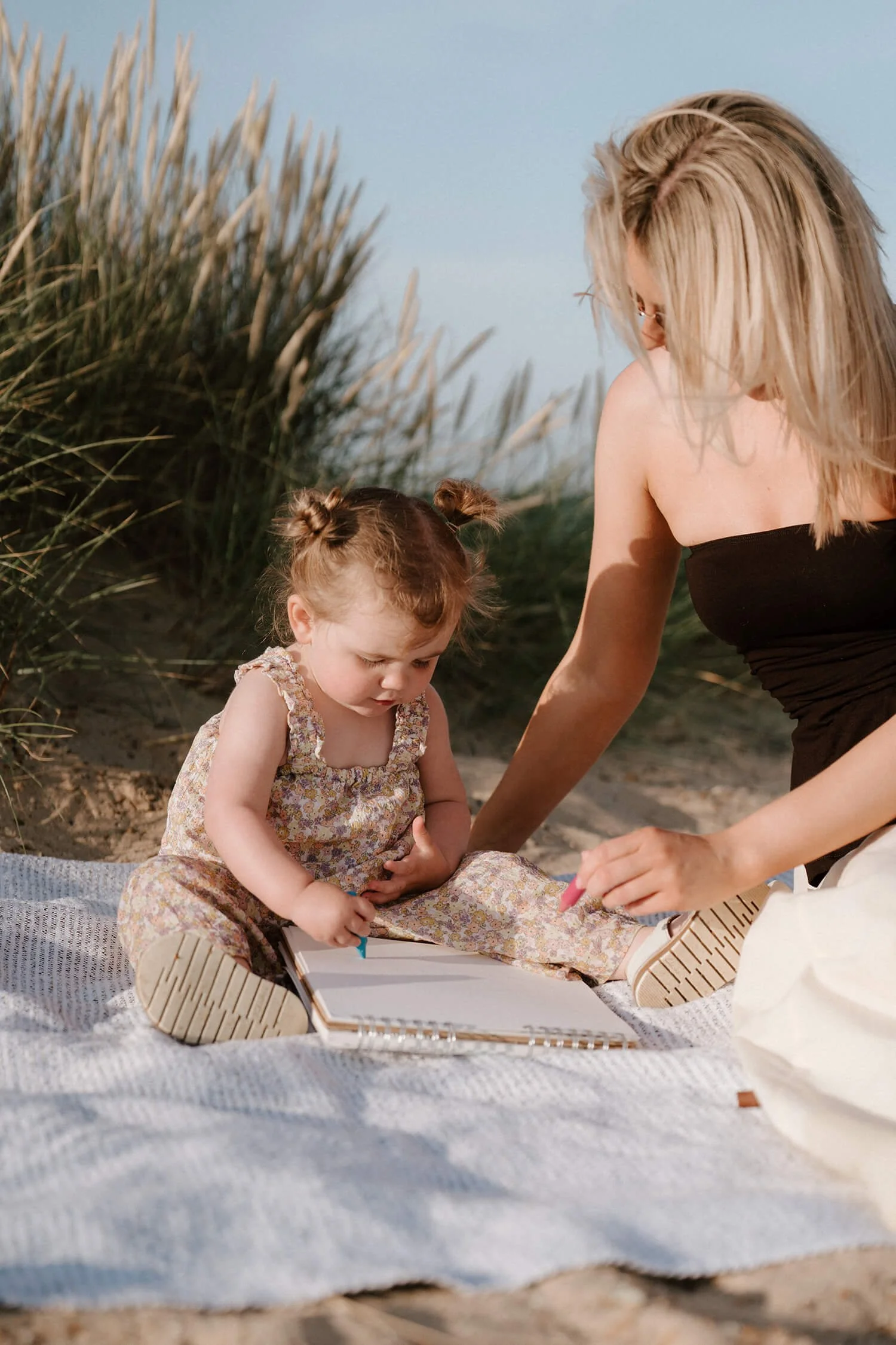 A woman and a young girl sitting on a blanket at the beach, looking at an open notebook.