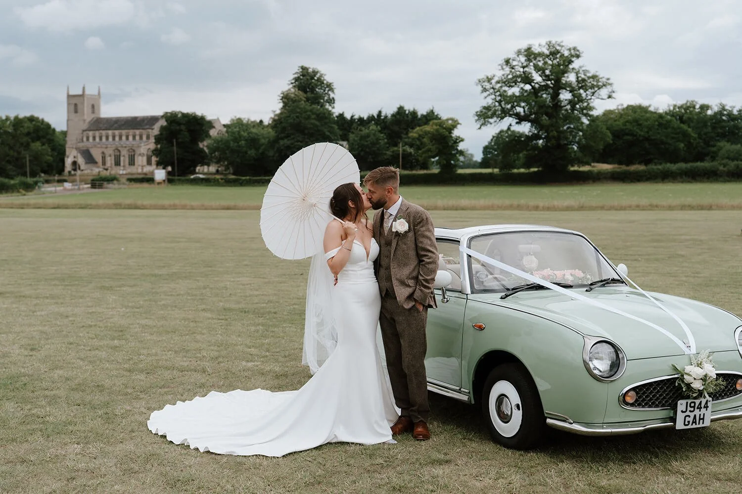 A bride and groom sharing a kiss, standing next to a vintage green car decorated with white ribbons and flowers, outdoors on a grassy field with a church and trees in the background.