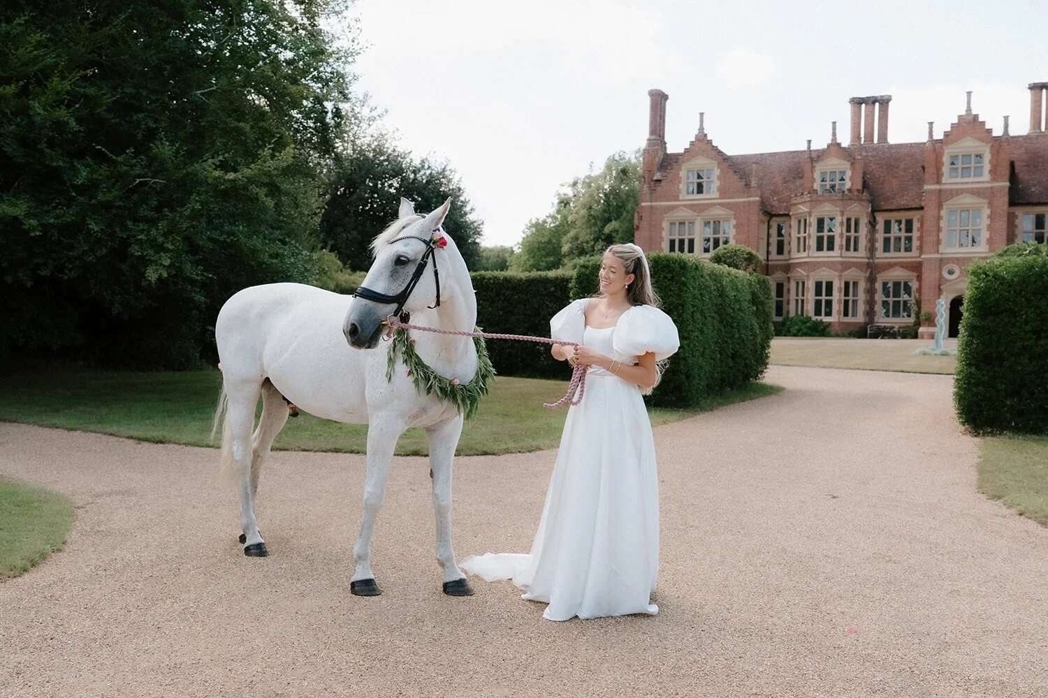 A woman in a white dress holding a pink rope leads a white horse decorated with a green leafy wreath in front of a large old brick house with multiple chimneys and windows, on a gravel pathway with green shrubs and trees.