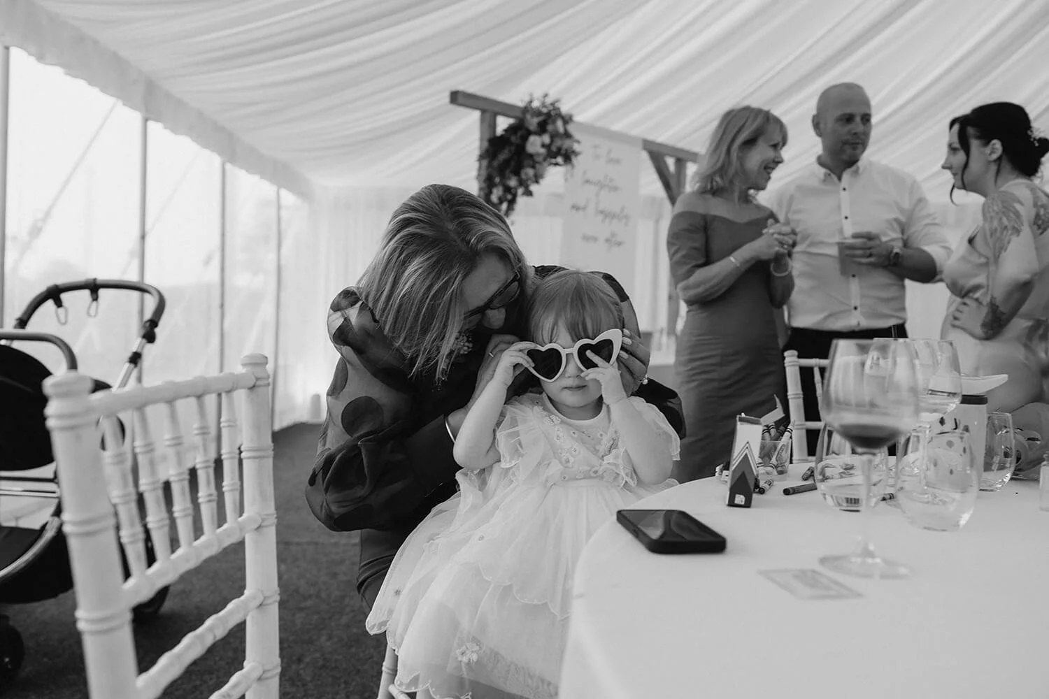 A young girl in a white dress and sunglasses sitting at a table with an adult woman leaning close to her, inside a decorated event tent, with several adults standing and conversing in the background.