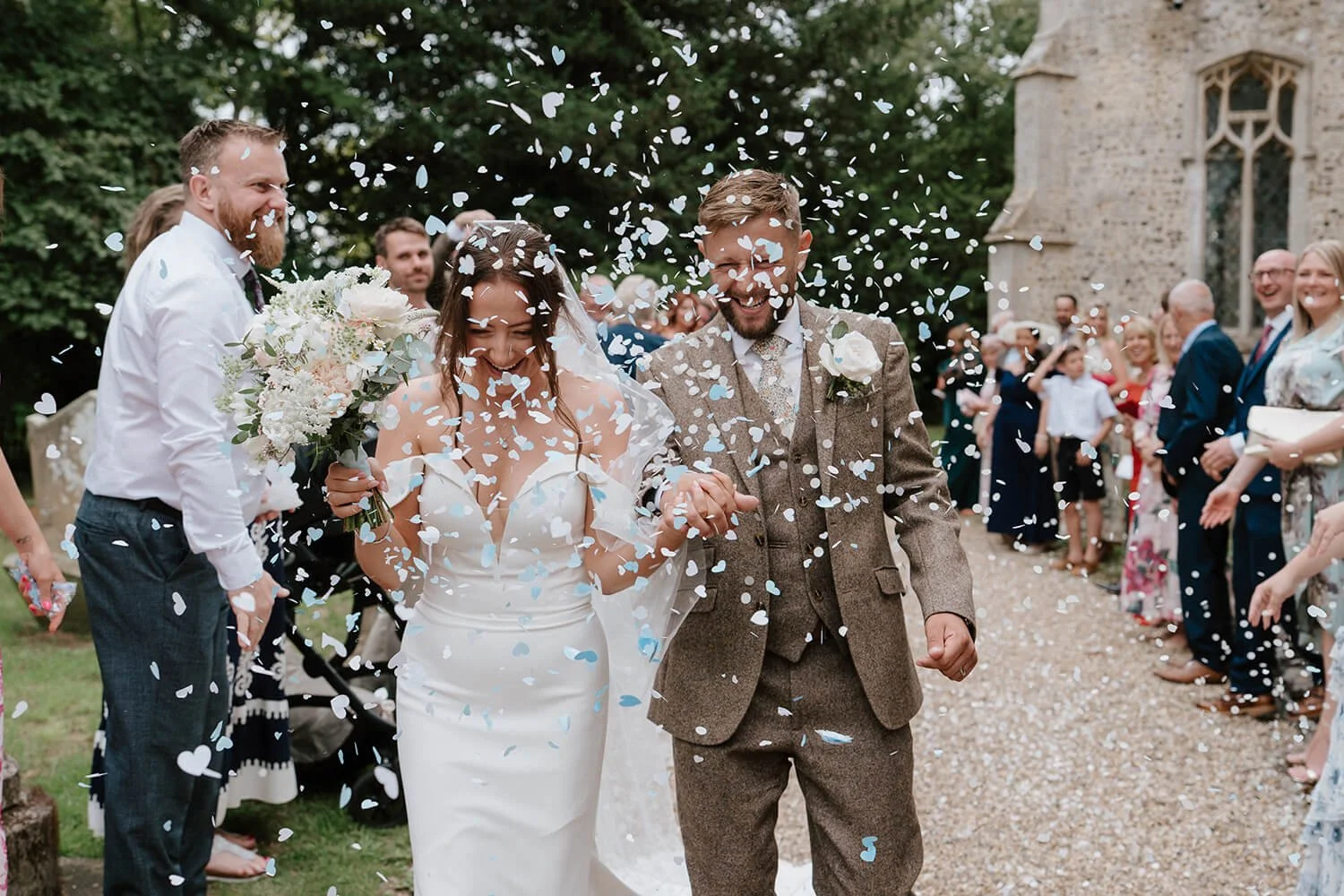 A newlywed couple walks hand in hand through a shower of white and blue confetti, surrounded by friends and family outside a stone church.
