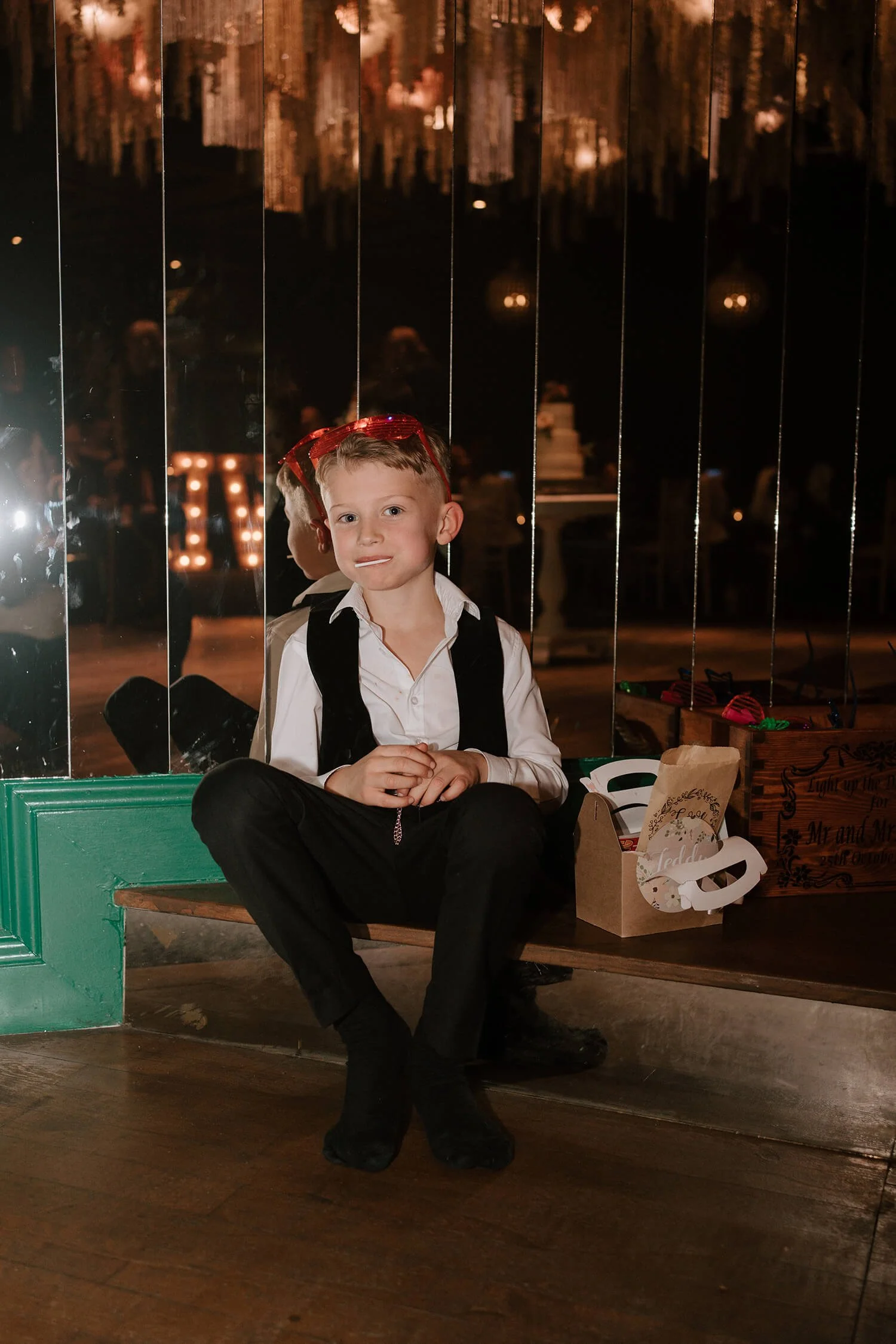 A young boy in formal attire sitting on a wooden bench at a party or celebration, with a backdrop of a reflective mirror and a dimly lit venue decorated with hanging lights and items on the bench nearby.