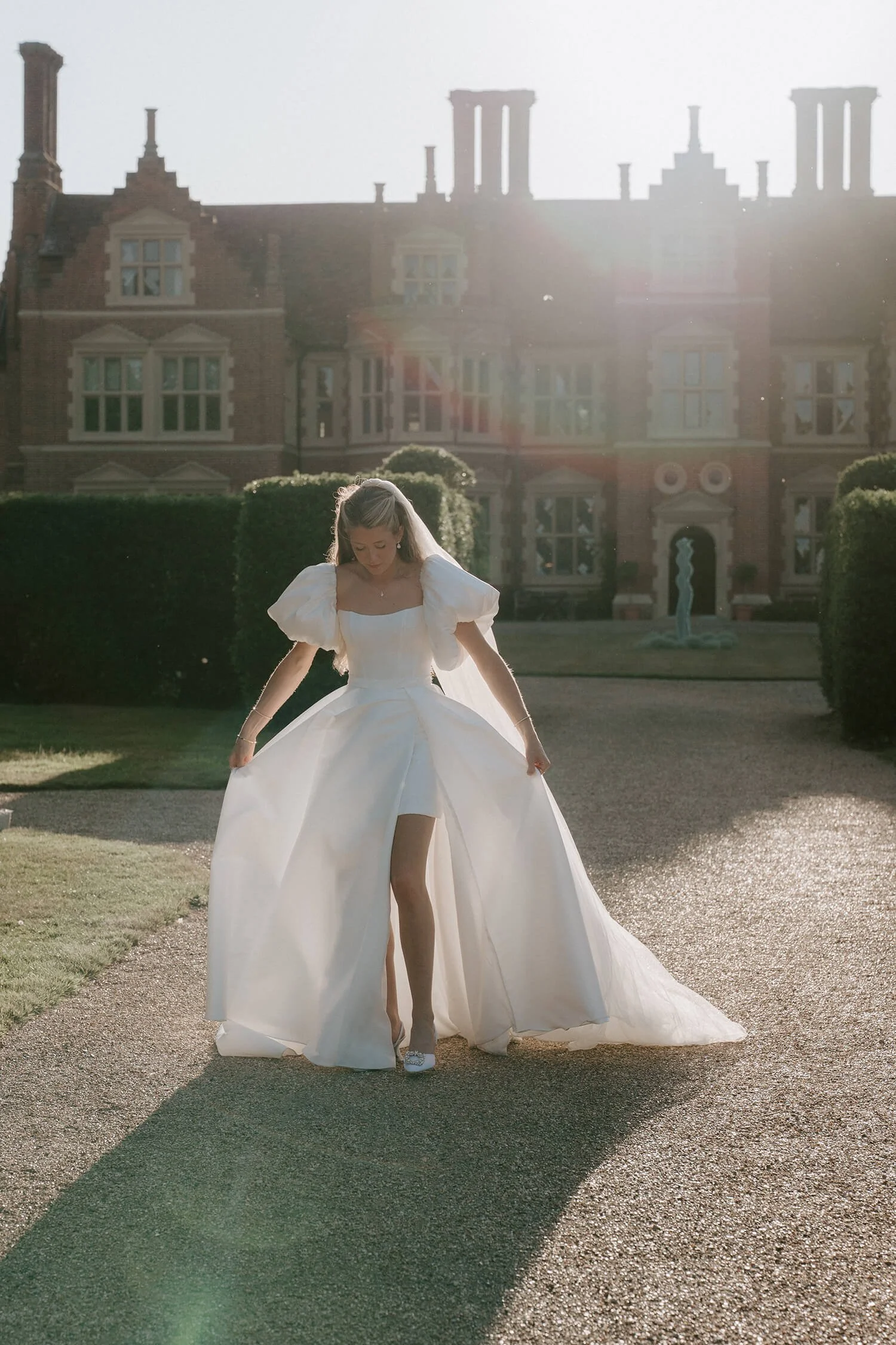 A bride in a white wedding dress standing on a driveway in front of a large historic mansion, with sunlight creating a halo effect behind her.