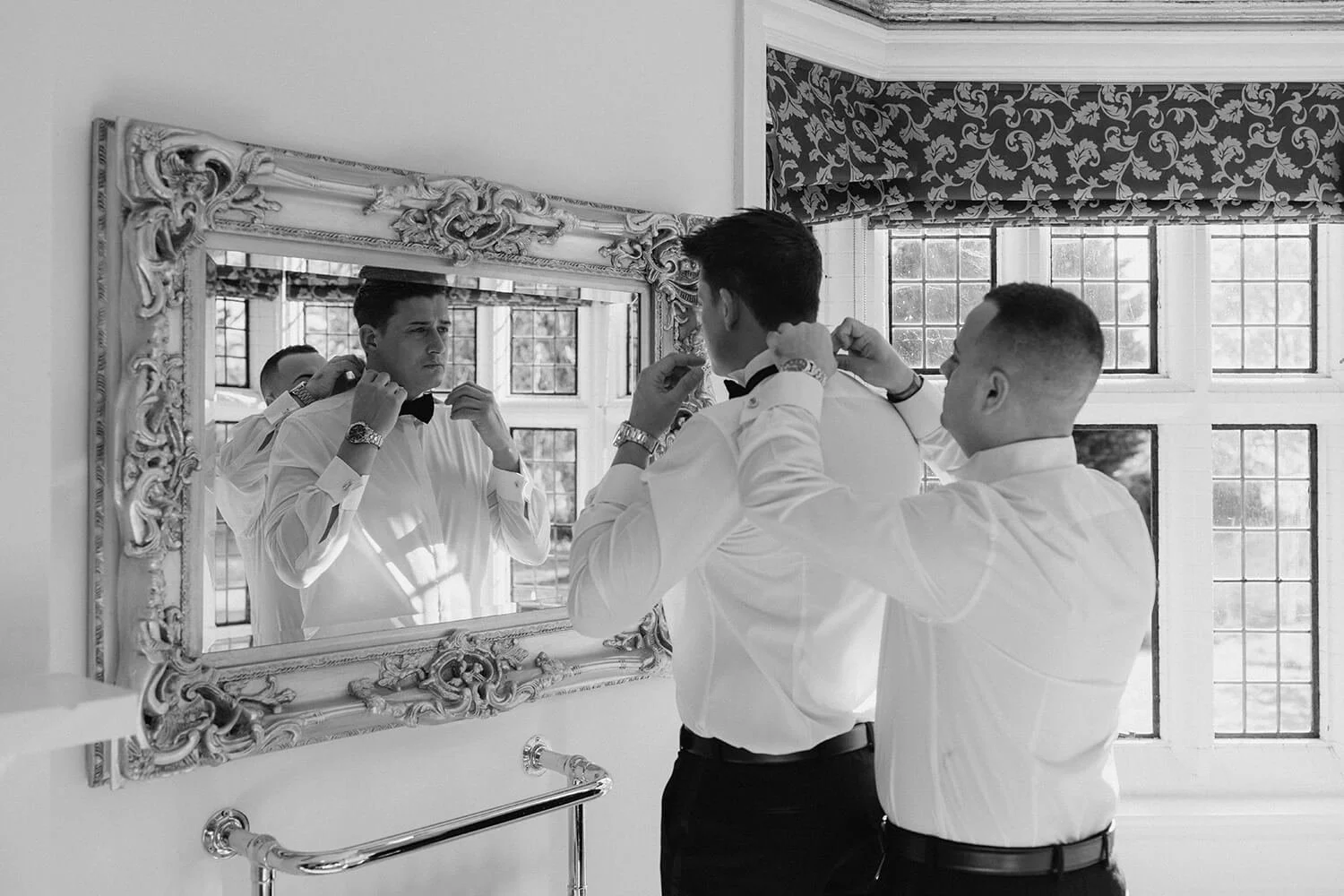 Two men are getting ready, adjusting their bow ties in front of a mirror. One man is looking at himself in the mirror while the other helps him. They are dressed in white shirts, with a large window in the background.