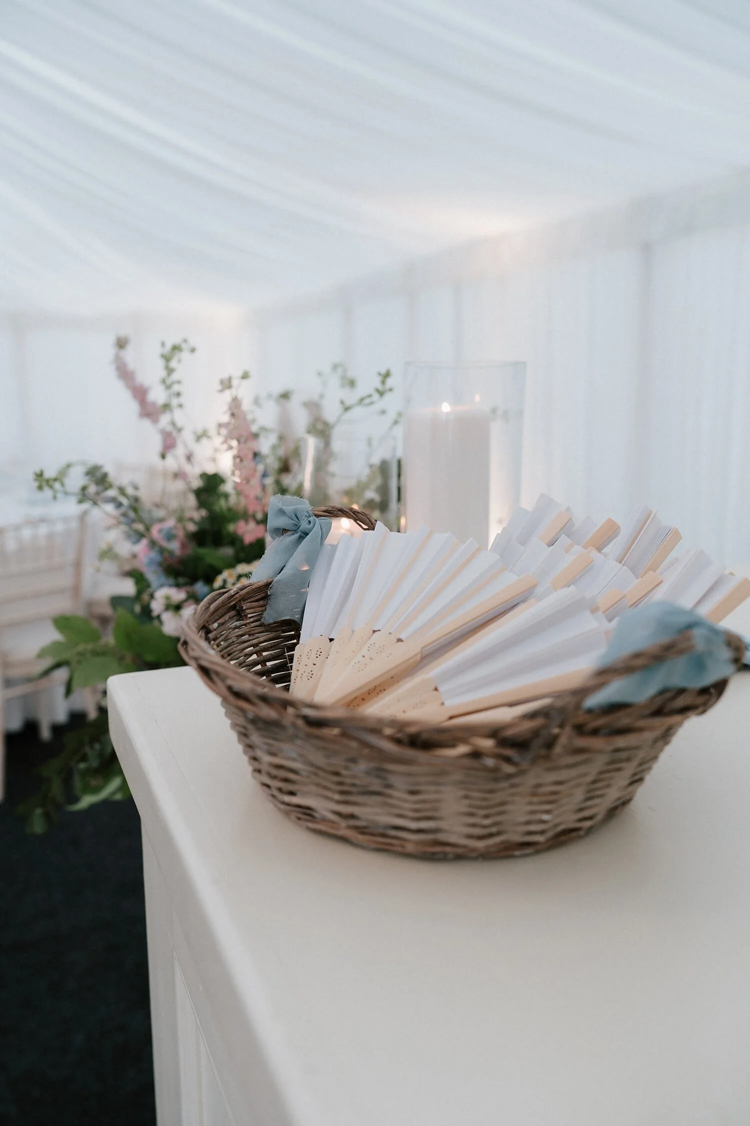 A wicker basket filled with white and light blue paper fans on a white table, with floral arrangements and candles in the background.