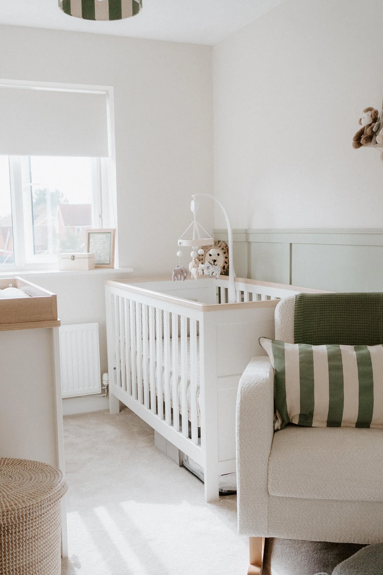 A cozy nursery with a white crib, a window with a white blinds and a framed photo on the windowsill, a beige armchair with a green and white striped pillow, and soft natural light.