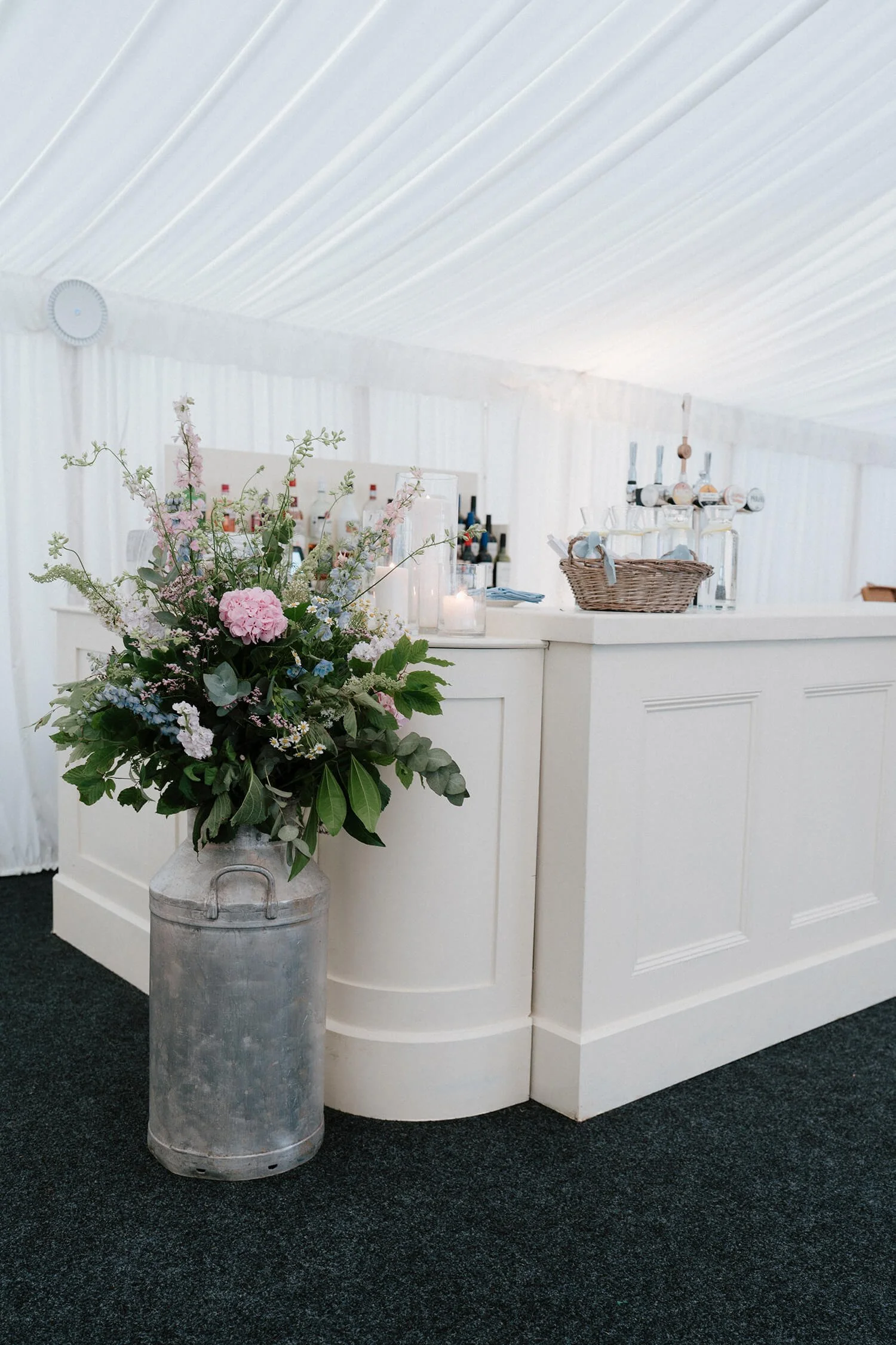 A white bar with a flower arrangement in a vintage milk can, bottles on shelves, and a basket with glasses inside a white tent at a wedding or event.