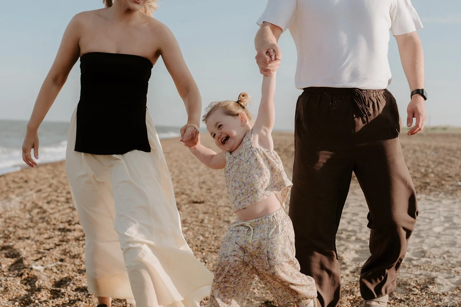 A young girl playing joyfully on the beach with her parents holding her hands, with the ocean in the background.