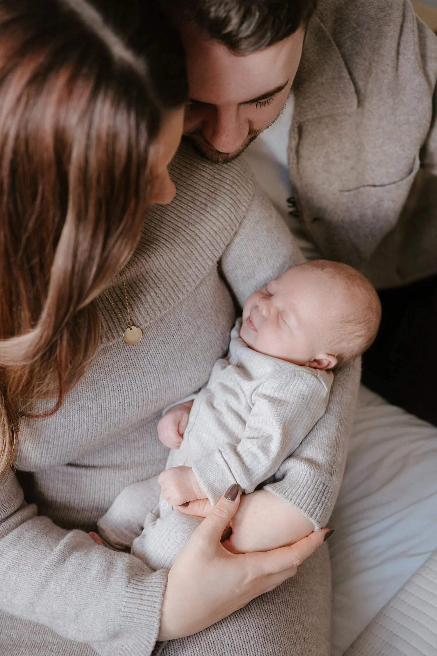 A family with a woman and man holding a newborn baby, who is smiling and dressed in beige, in a cozy indoor setting.