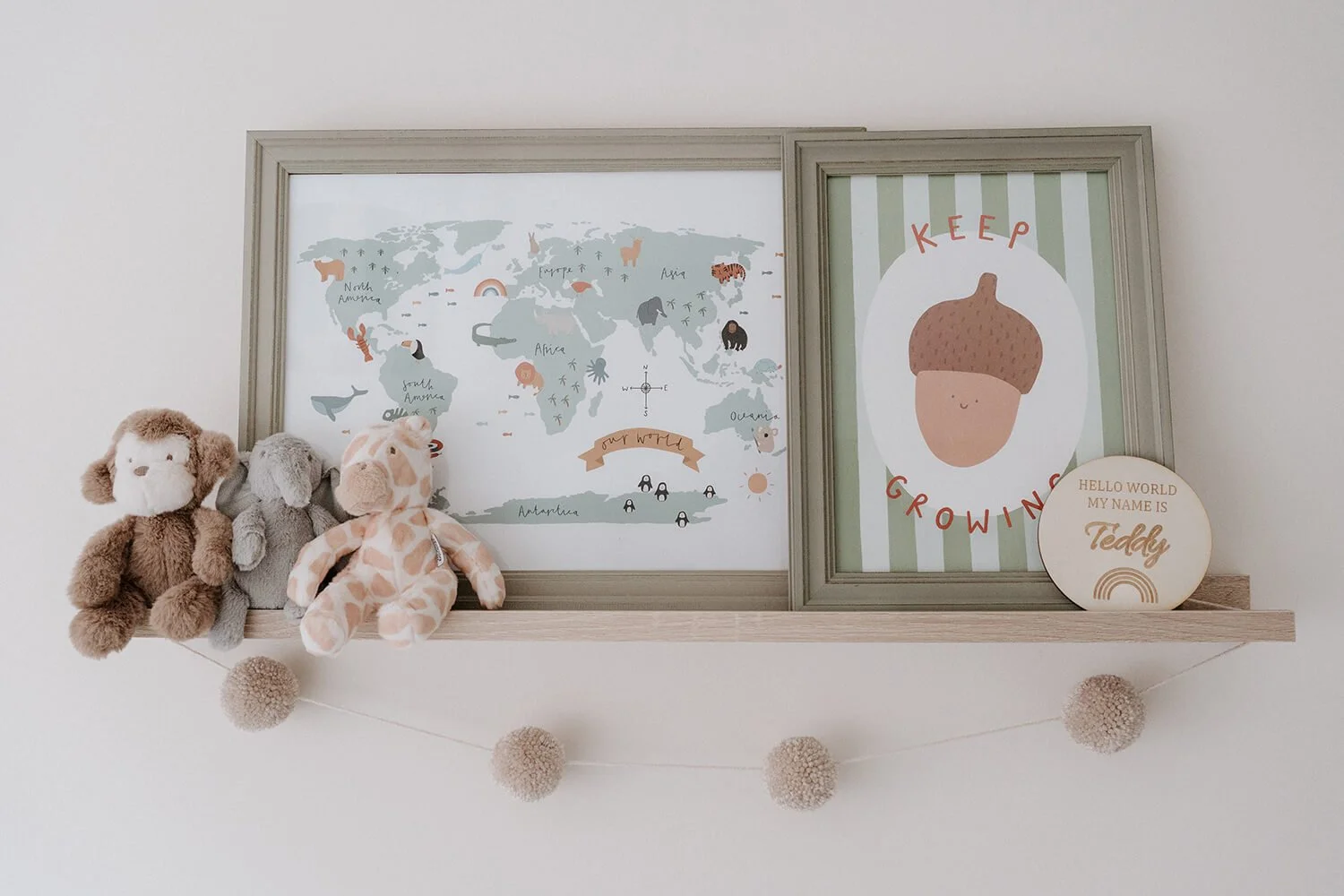 Decorative nursery shelf with plush toys, framed world map, baby growth chart, and a circular sign with the name Teddy, beneath a string of beige pom-pom garland.