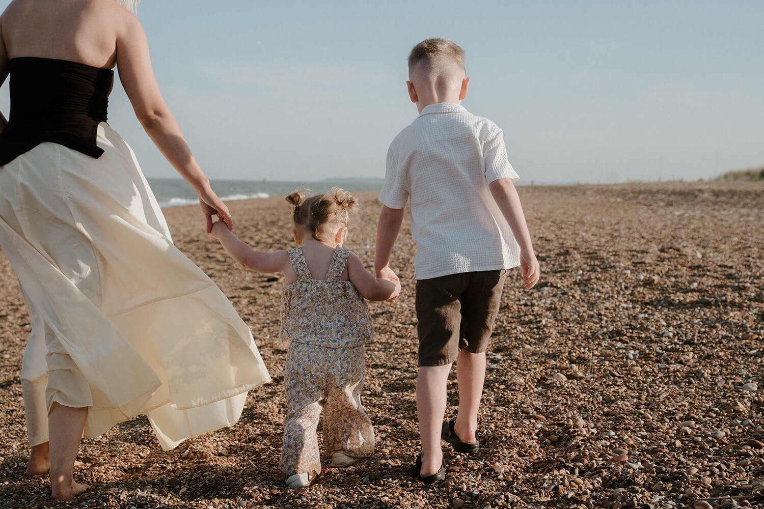 A woman with two children walking barefoot on a pebble beach, holding their hands, with the ocean in the background