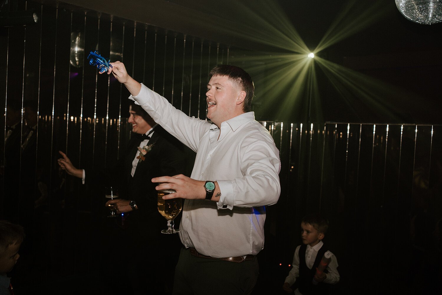 People celebrating at a party, with a man in a white shirt holding a champagne glass and dancing, and another man in a black suit holding a drink, illuminated by colorful lights.