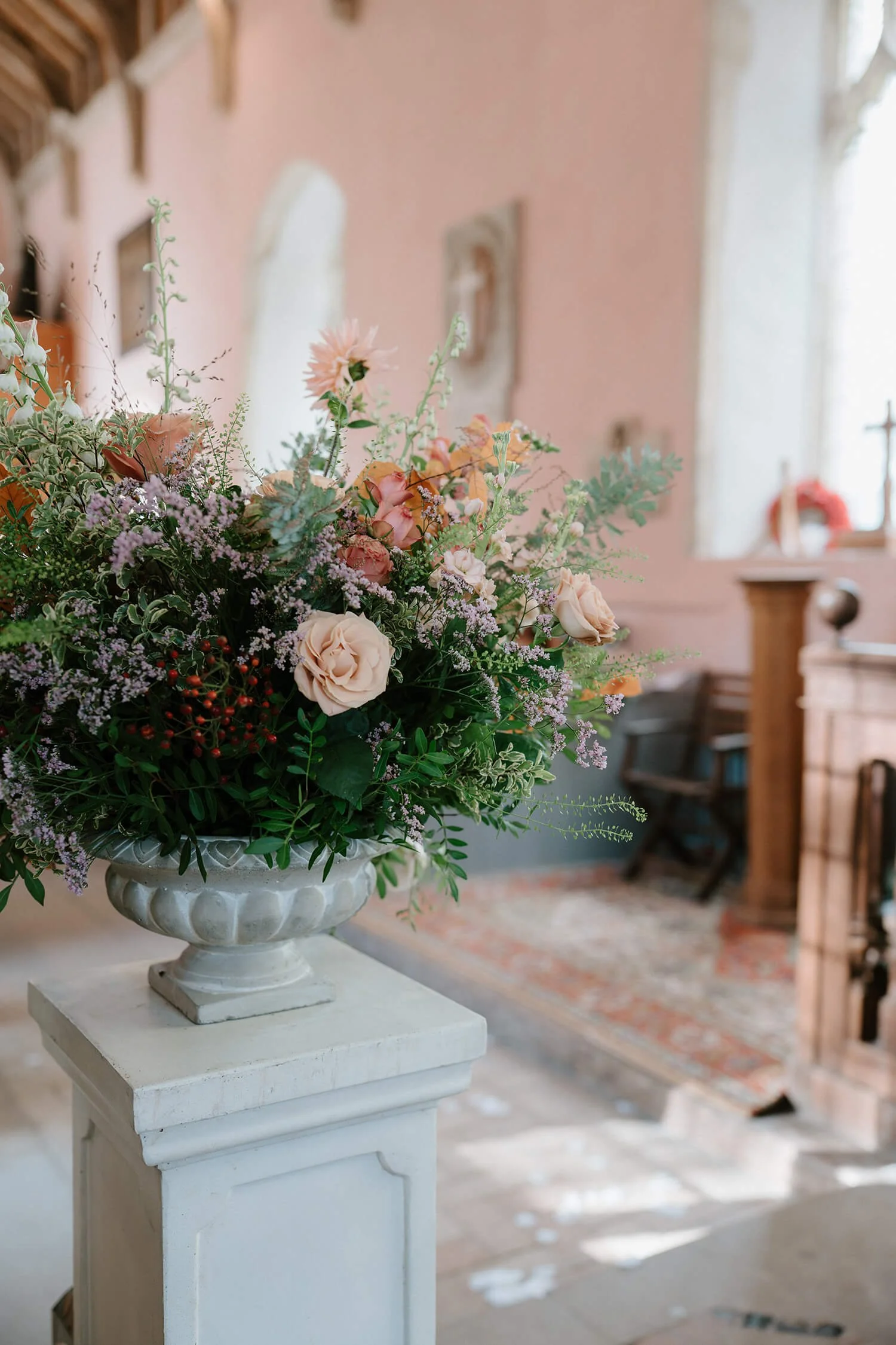 A floral arrangement with pink, peach, and purple flowers in a white urn on a pedestal inside a cozy, rustic room with pink walls and vintage decor.