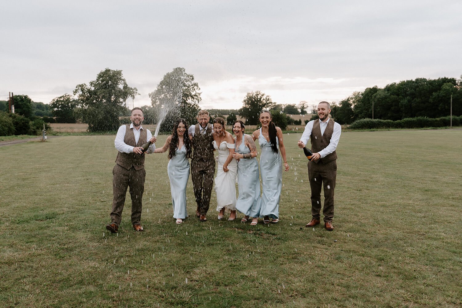 Group of seven people celebrating outdoors, spraying champagne and laughing, on a grassy field with trees and cloudy sky in the background.