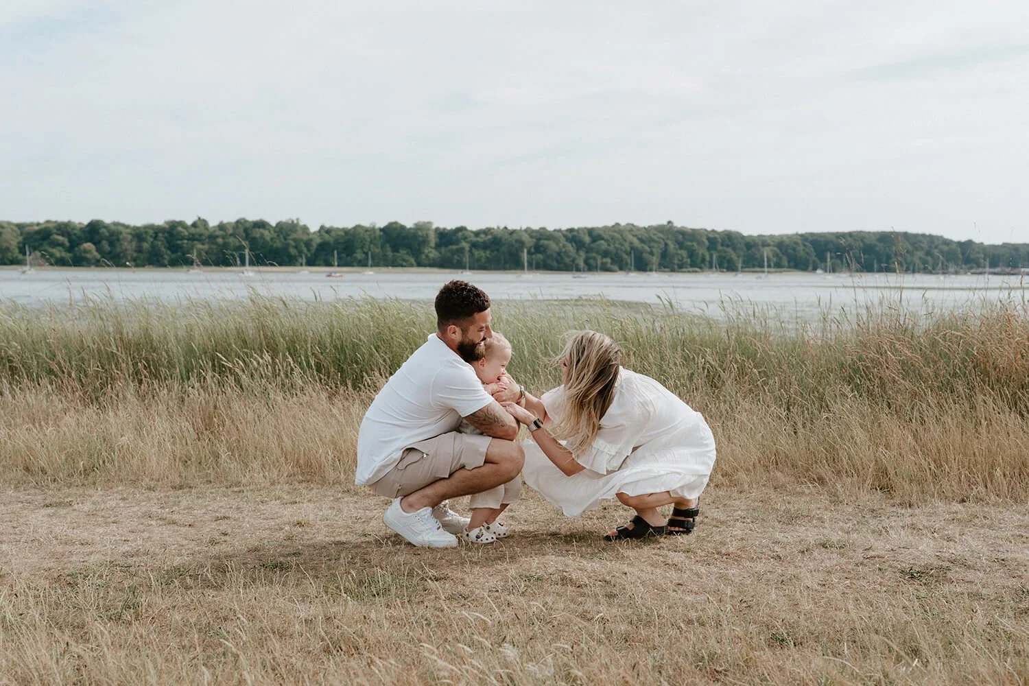 A family of three on a grassy lakeside, with a man and woman holding their toddler and sharing a tender moment, under a cloudy sky with trees in the background.