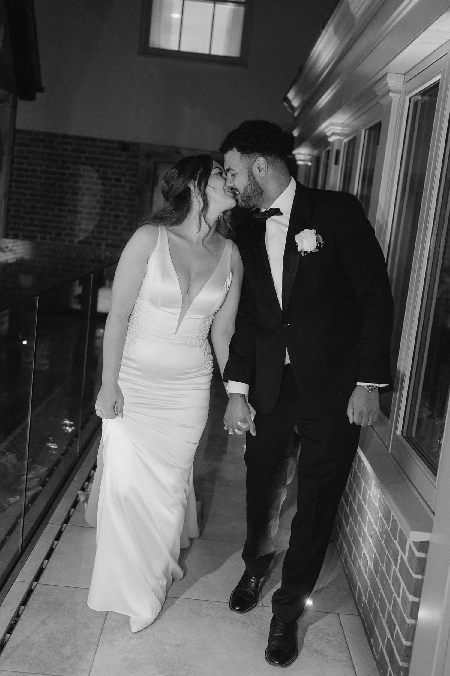 A black and white photo of a bride and groom holding hands and leaning in for a kiss on a balcony or porch, dressed in wedding attire, with windows and brick wall in background.