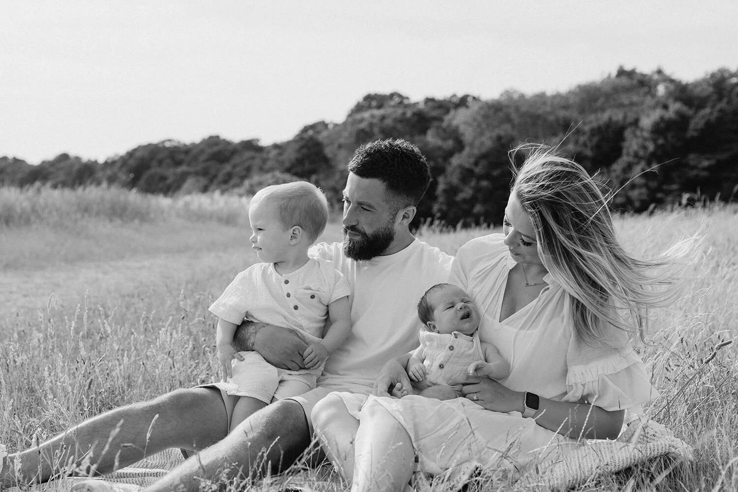 A black and white photo of a family sitting in a grassy field. The father is holding a toddler, and the mother is holding a newborn. The family is outdoors with trees in the background.