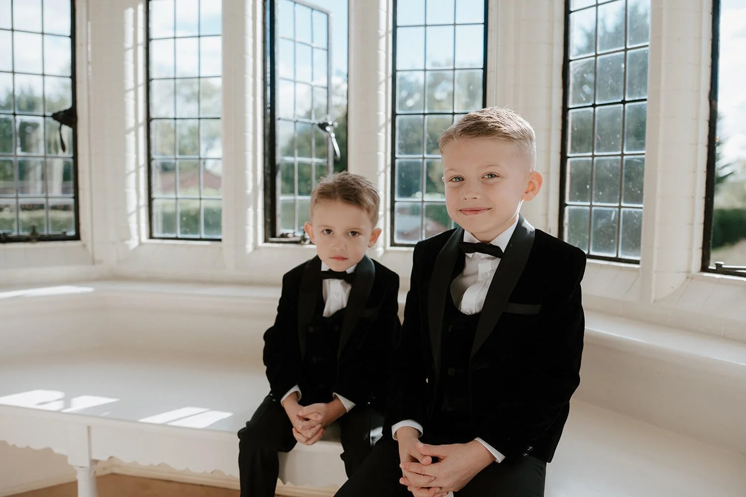 Two young boys dressed in black tuxedos with bow ties sitting inside a room with large grid windows and white walls, one smiling and one with a serious expression.
