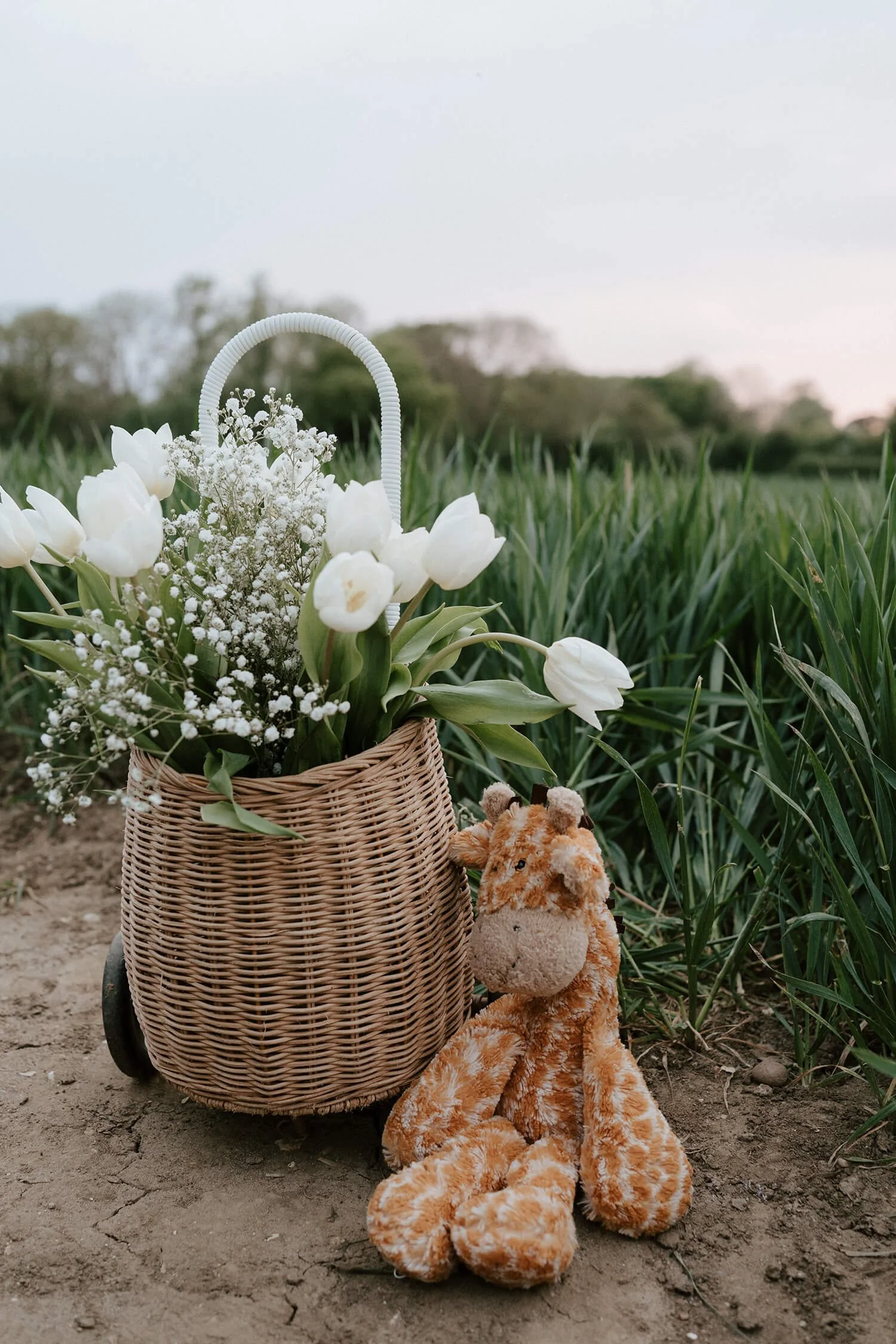 A woven basket filled with white tulips and baby's breath flowers, accompanied by a plush giraffe toy, arranged outdoors with grass and trees in the background.