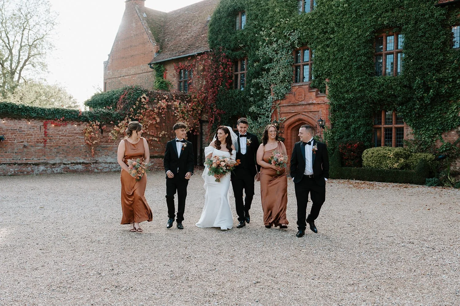 A wedding party walking together outside in front of a brick building covered with green and red ivy. The group includes a bride in a white wedding dress with a floral bouquet, a groom in a black tuxedo, and four attendants dressed in formal attire, 