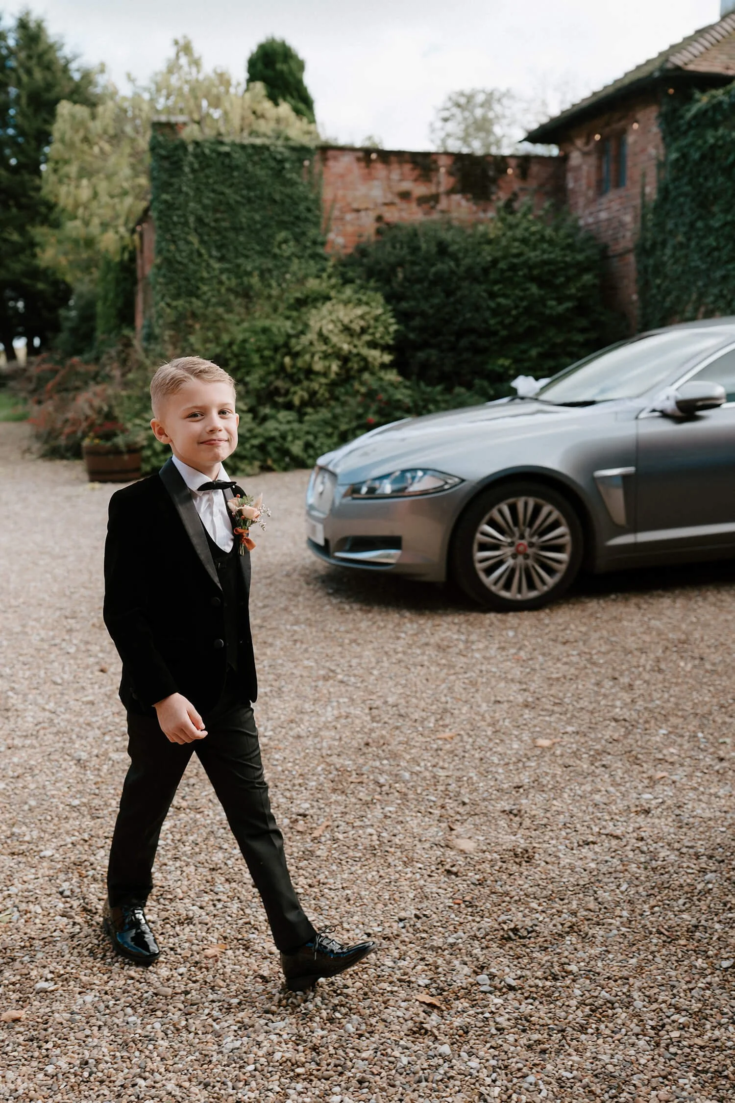 A young boy dressed in a black tuxedo with a boutonniere, walking on a gravel driveway next to a decorated silver car, with a background of greenery and a brick house.