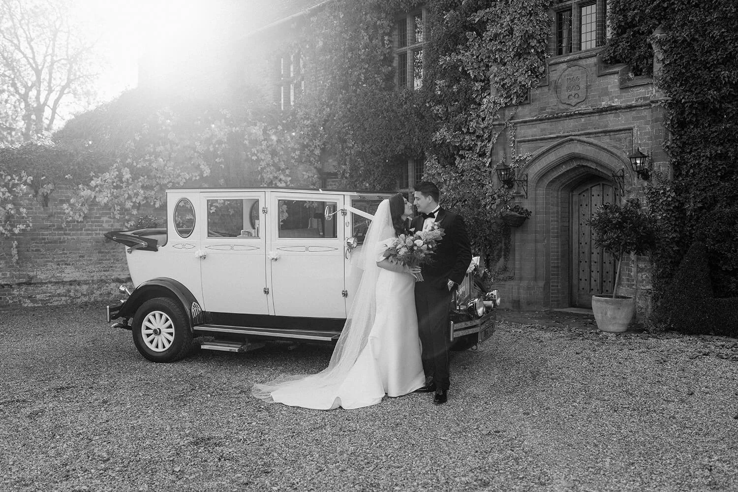 A bride and groom in wedding attire sharing a kiss next to a vintage car outside a historic building with ivy-covered walls.
